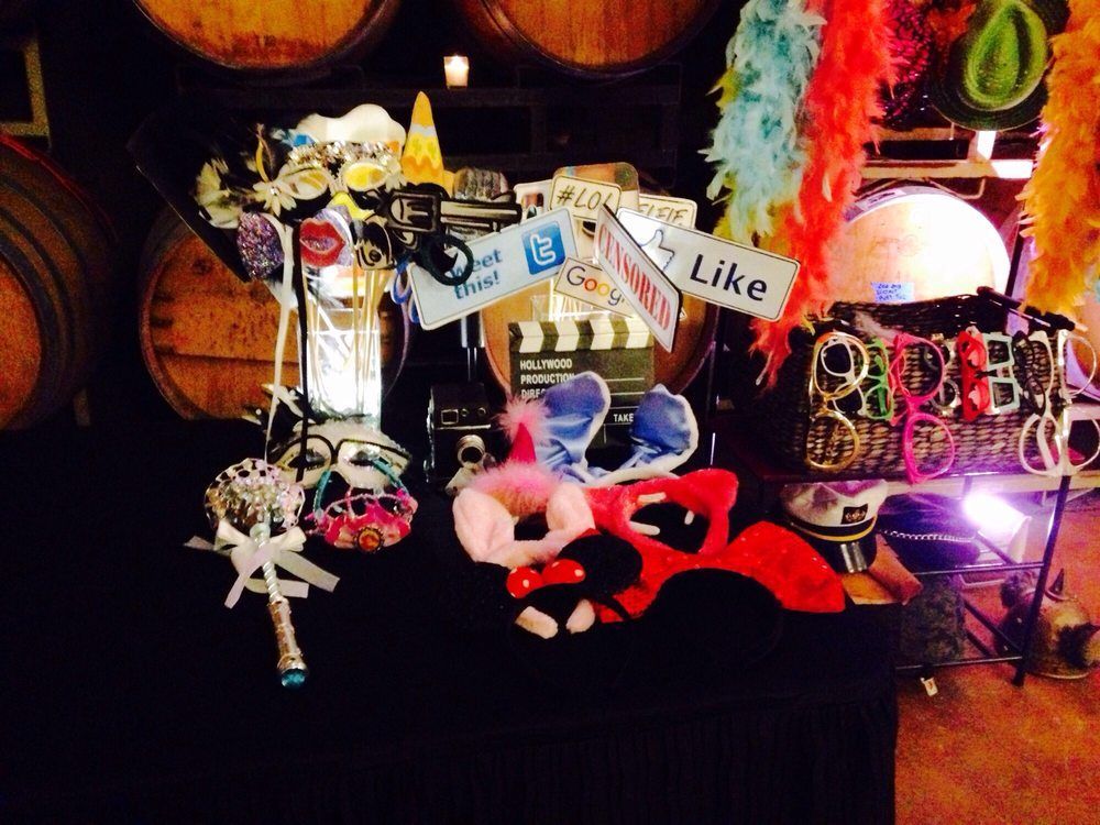 Photo booth props on a table: feather boas, masks, social media signs, glasses, and a bouquet. Wine barrels in background.