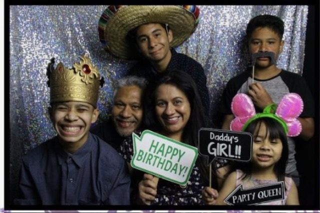 Family photo booth portrait, silver backdrop. People hold props like a crown, sombrero, and signs. Happy expressions.