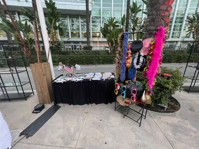 A table and stand with various colorful hats, sunglasses, and boas outside.