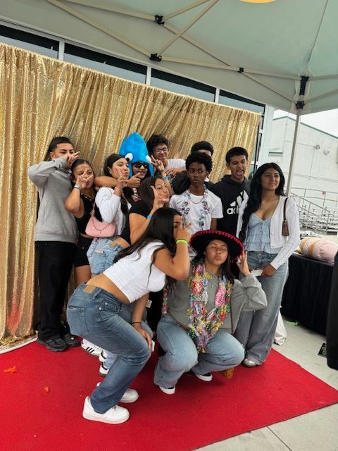 Group of people posing on a red carpet in front of a gold backdrop. They are making silly gestures and faces.