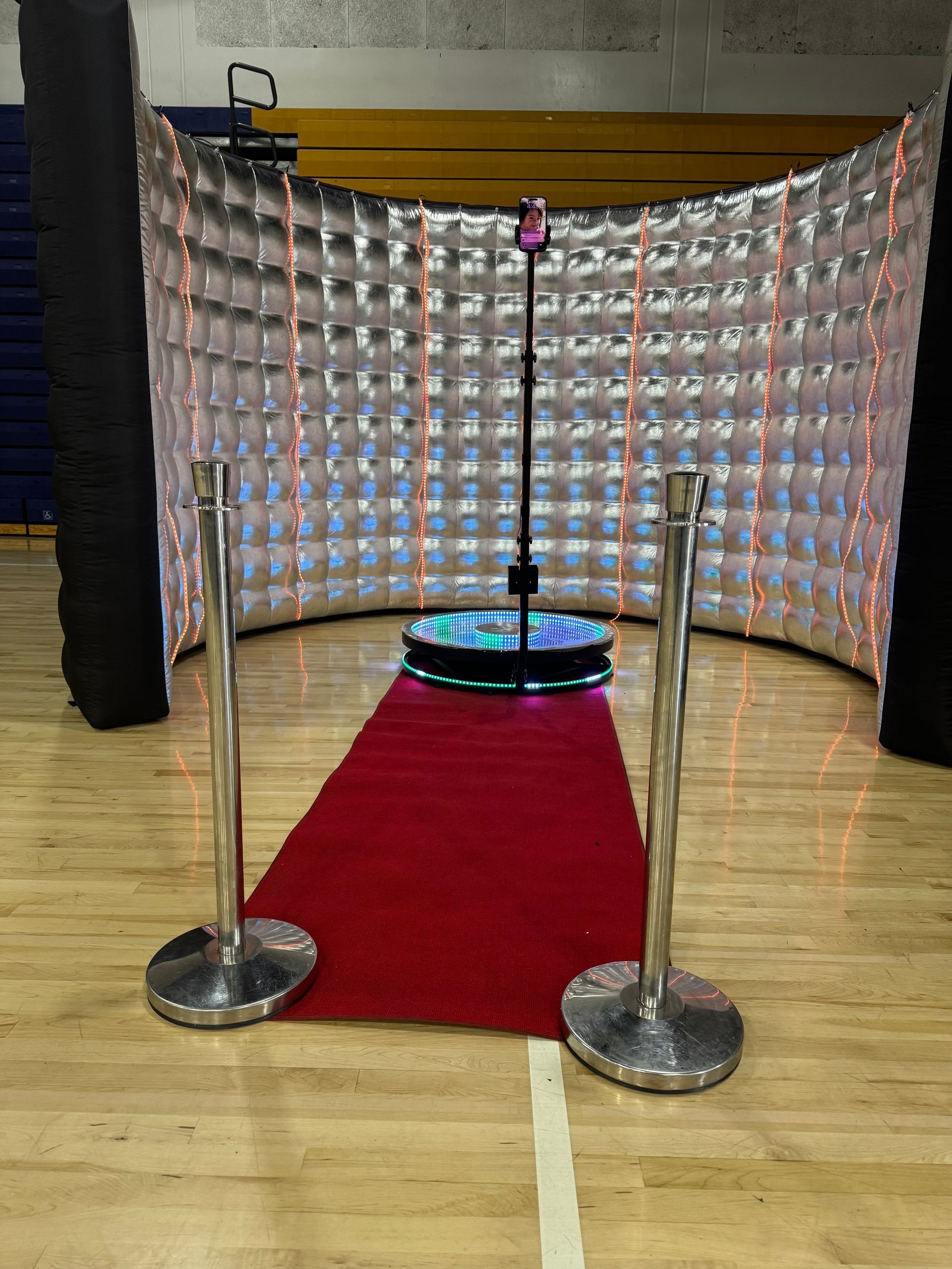 360 photo booth setup with red carpet, stanchions, silver backdrop, and camera on a stand.