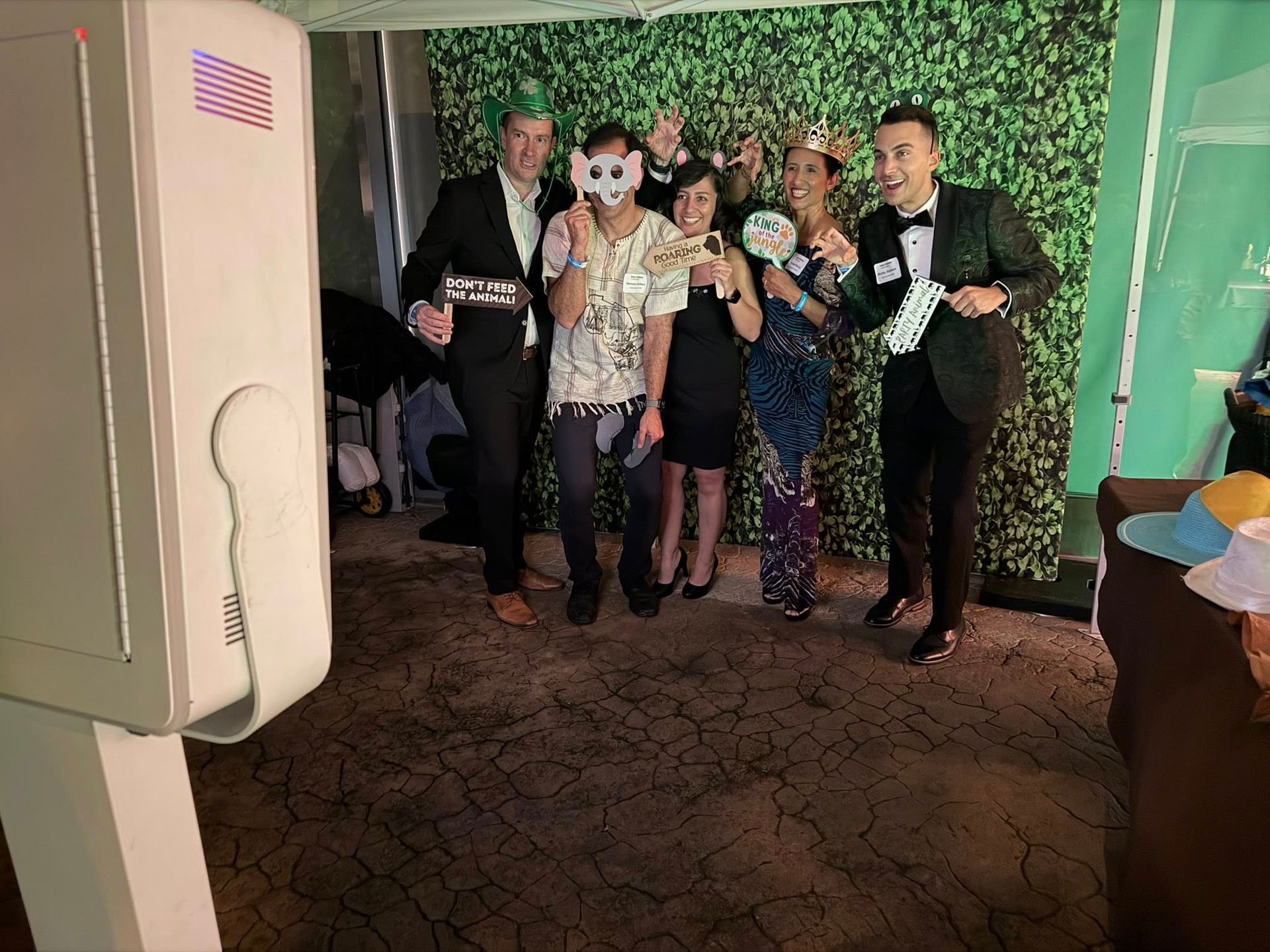 People posing with props in a photo booth at an event. Green backdrop, brown carpet.