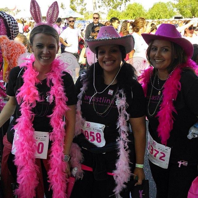 Three women in pink and black outfits at a breast cancer awareness walk, wearing boas and hats.