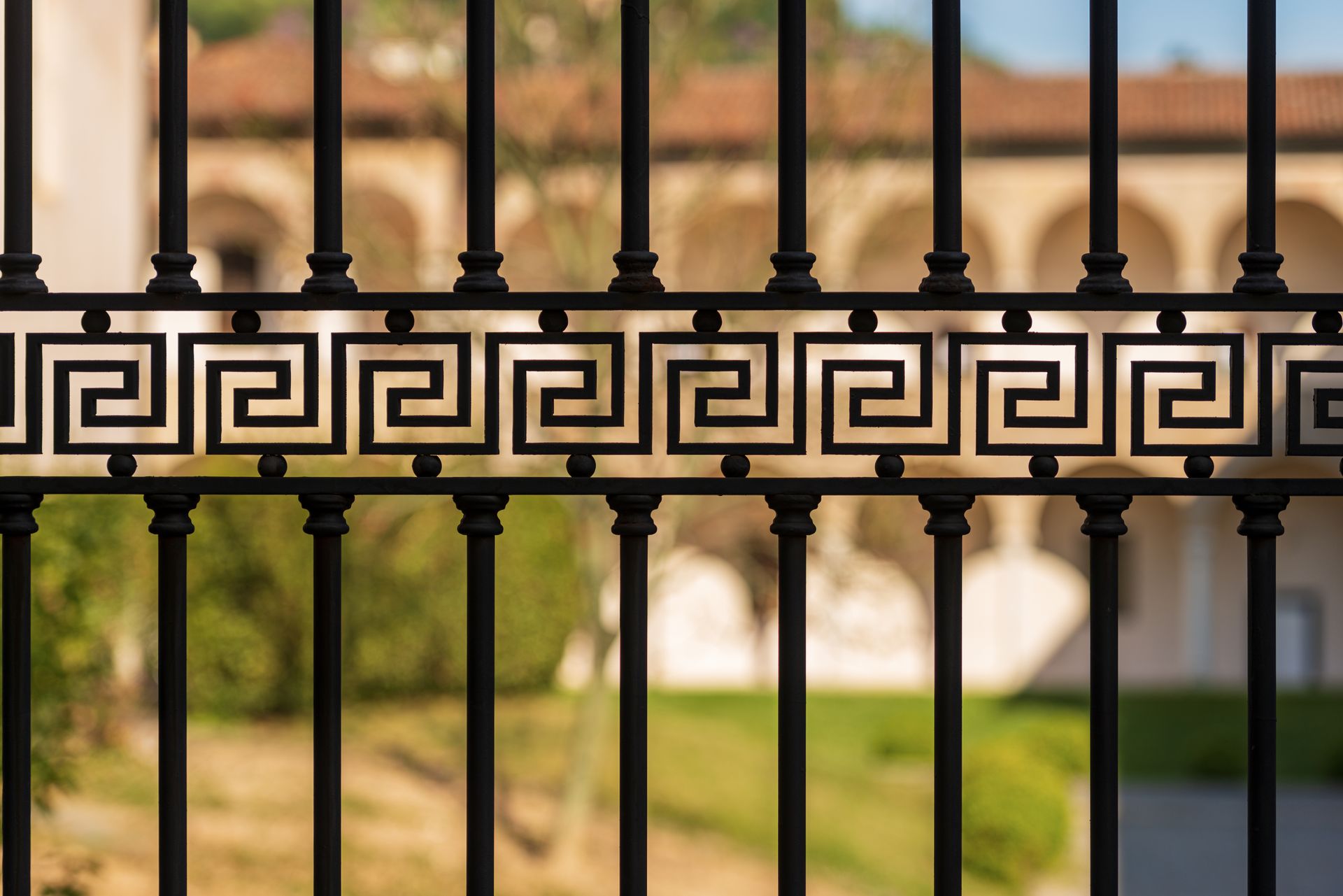 Recinzione in ferro nero con motivo a greca, sfondo sfocato di un edificio e fogliame.