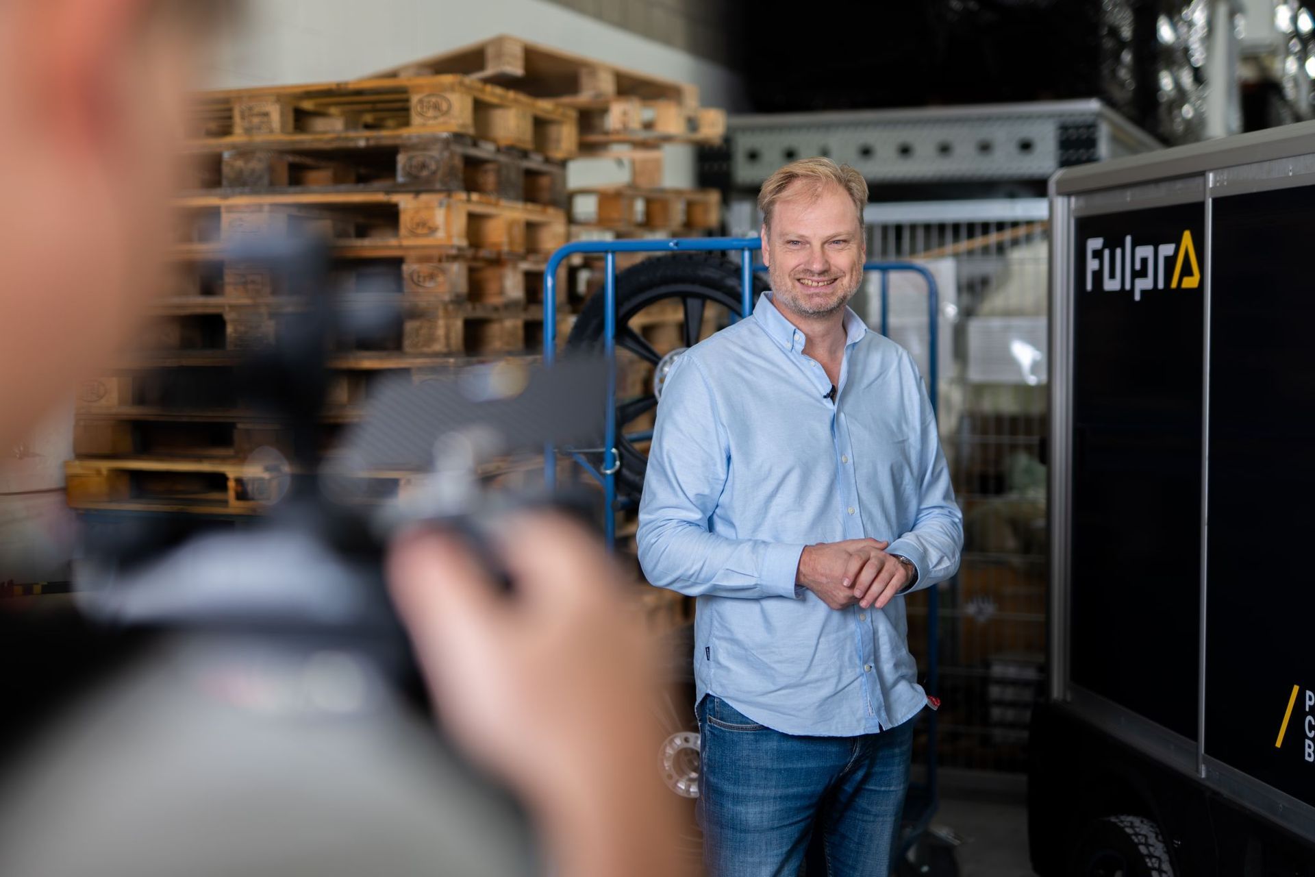 Een man in een blauw shirt en spijkerbroek lacht in een magazijn. Op de voorgrond is filmapparatuur te zien. Op een voertuig staat 