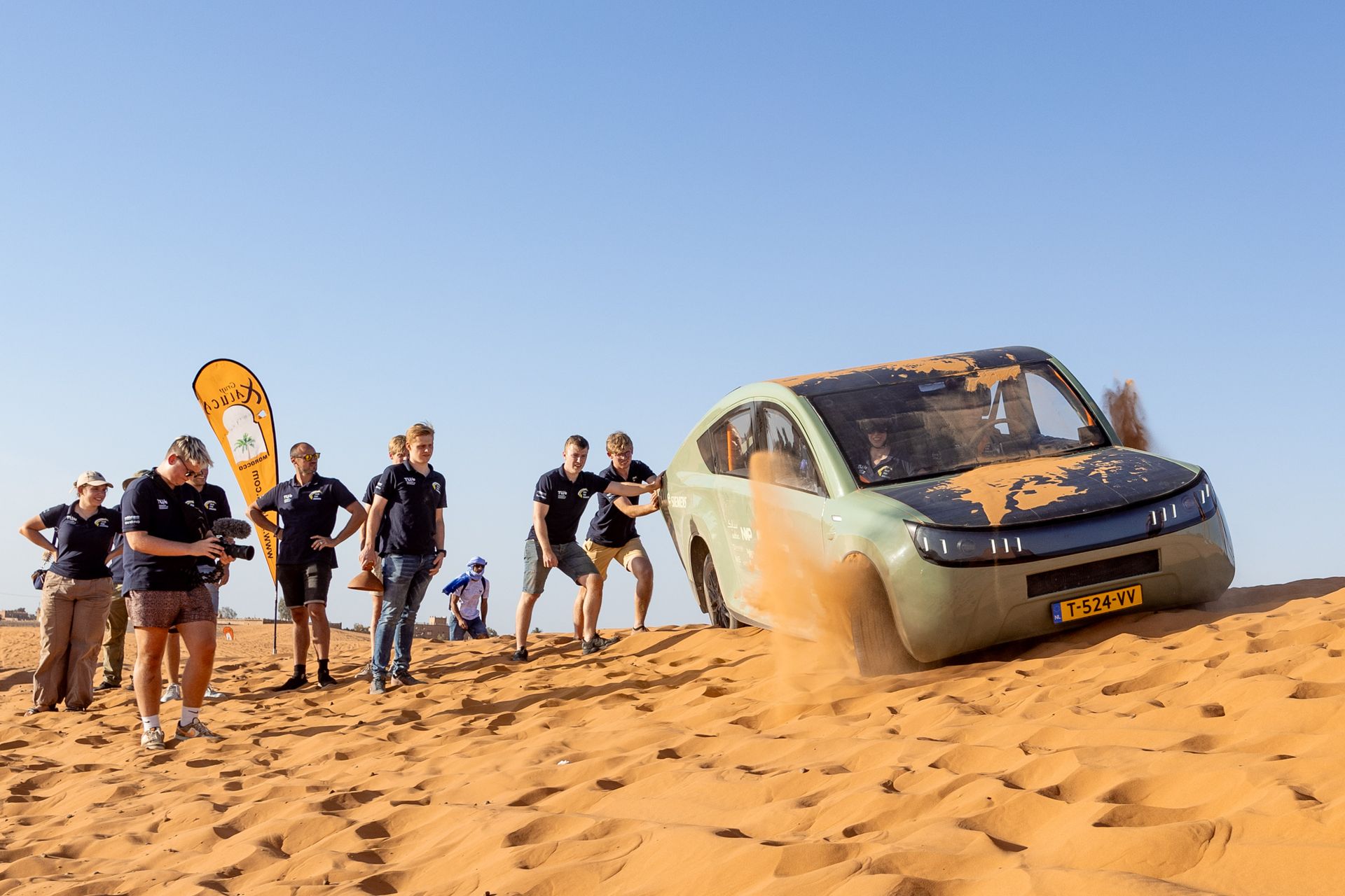 Een auto op zonne-energie zit vast in het zand; teamleden duwen hem door de woestijn.