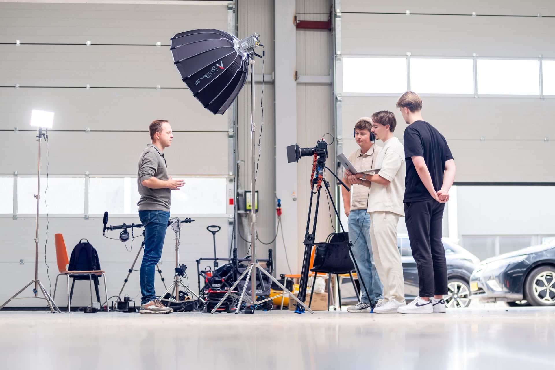 Een man wordt gefilmd in een garage door een filmploeg; een camera op statief, verlichtingsapparatuur en auto's zijn zichtbaar.