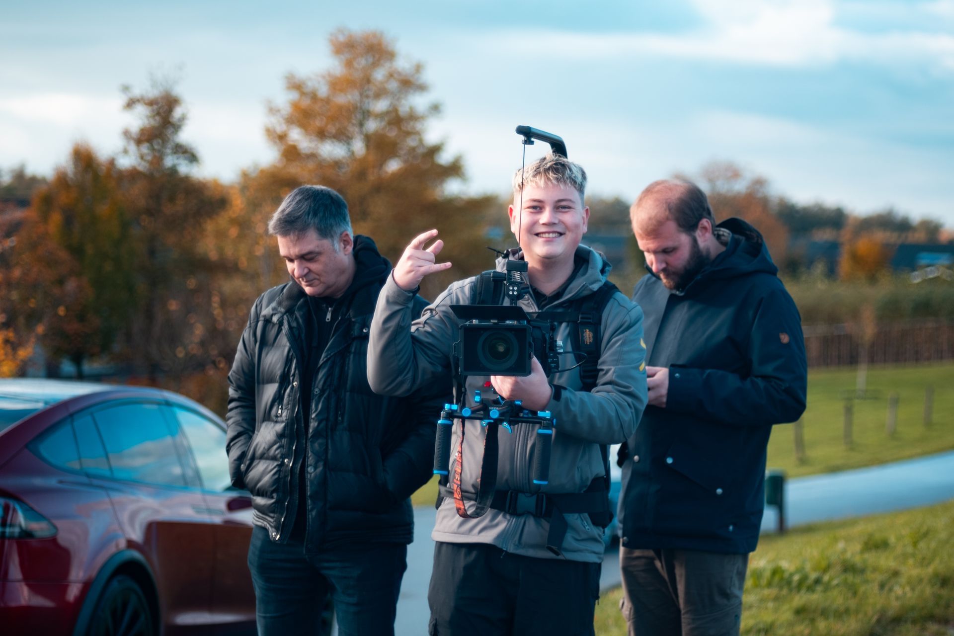 Drie mensen staan ​​buiten, één houdt een camera-opstelling vast en gebaart, de anderen kijken toe. Herfstachtergrond met een rode auto.