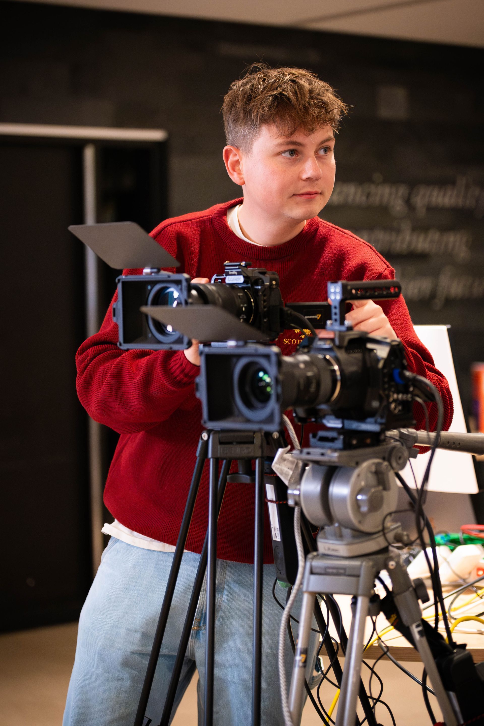 Een persoon in een rode trui bedient een videocamera op een statief. Het lijkt een binnenruimte te zijn.