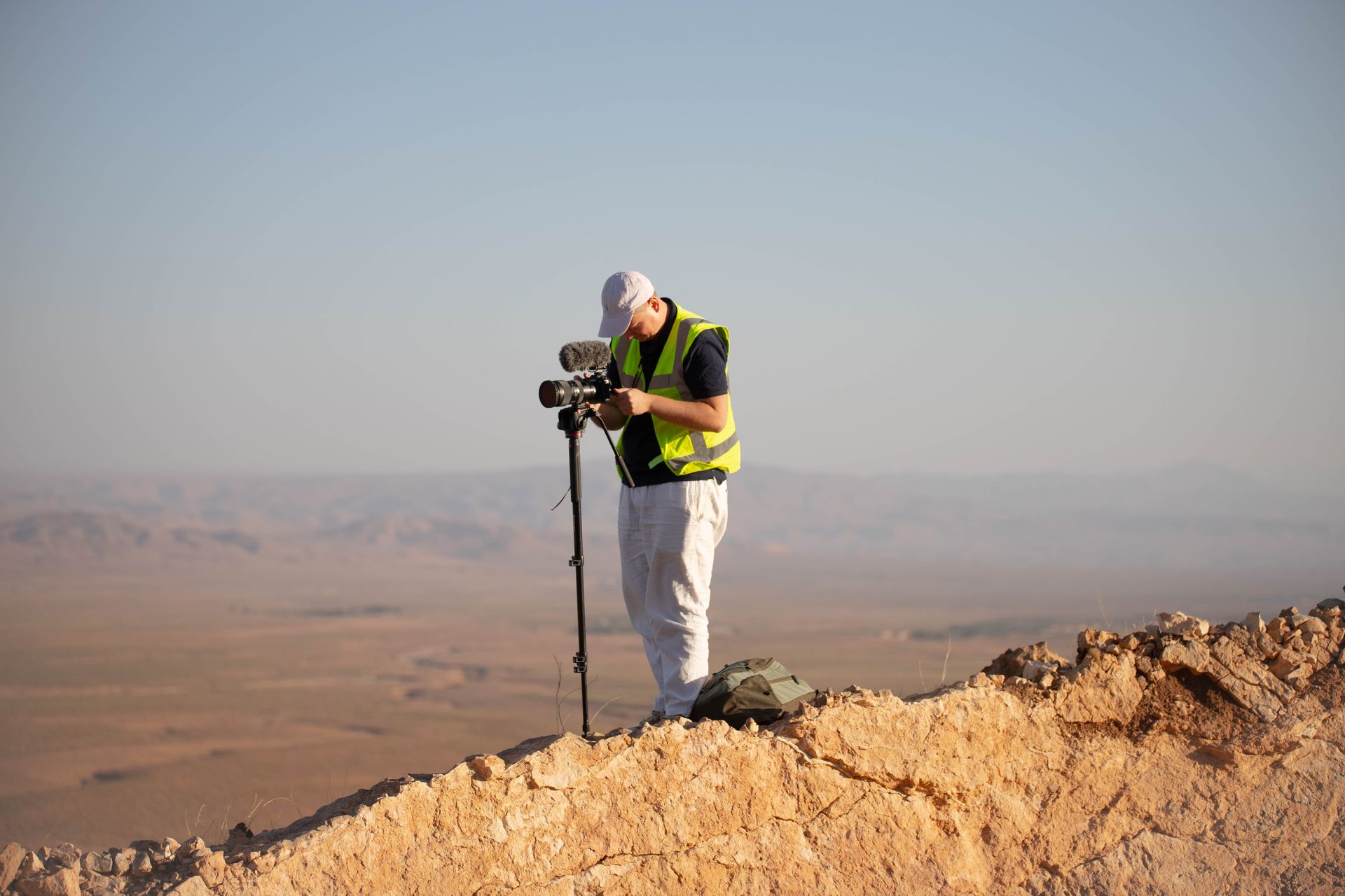 Een man, gekleed in een geel hesje, filmt een landschap met een camera op een statief vanaf een woestijnklif.