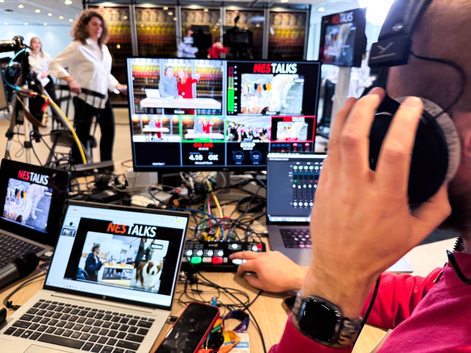 Een persoon met een koptelefoon bedient videoapparatuur. Op meerdere schermen is een live-uitzending te zien.