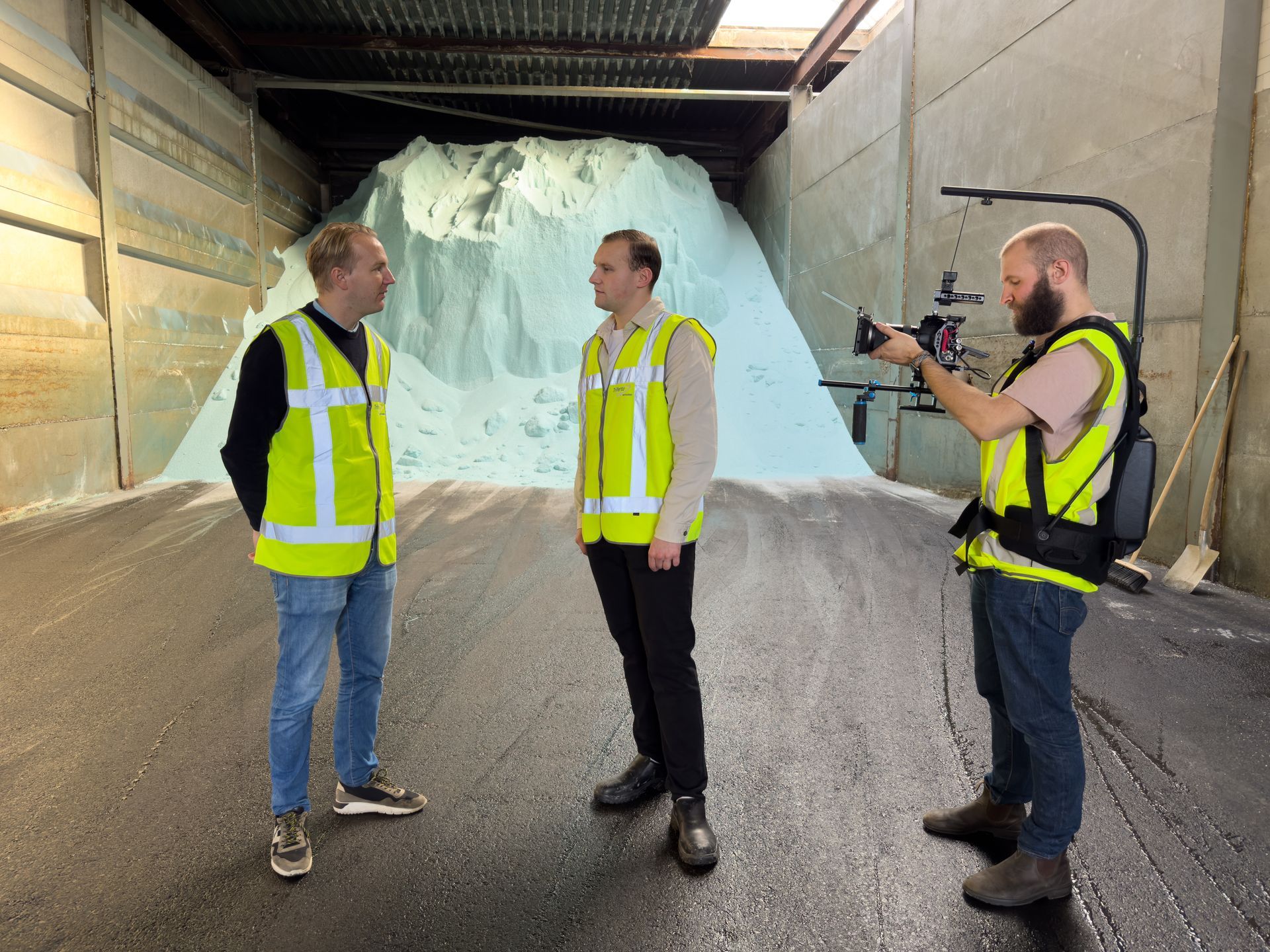 Drie mannen in reflecterende hesjes. Een van hen filmt twee mannen die bij een grote zouthoop in een magazijn staan.