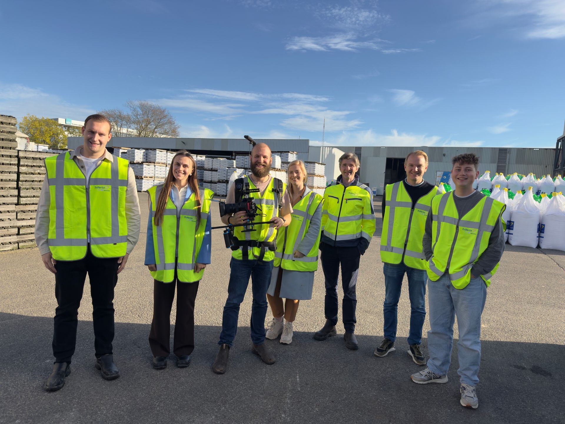 Een groep van zeven mensen, gekleed in veiligheidsvesten, staat buiten, in de buurt van betonblokken en materialen.
