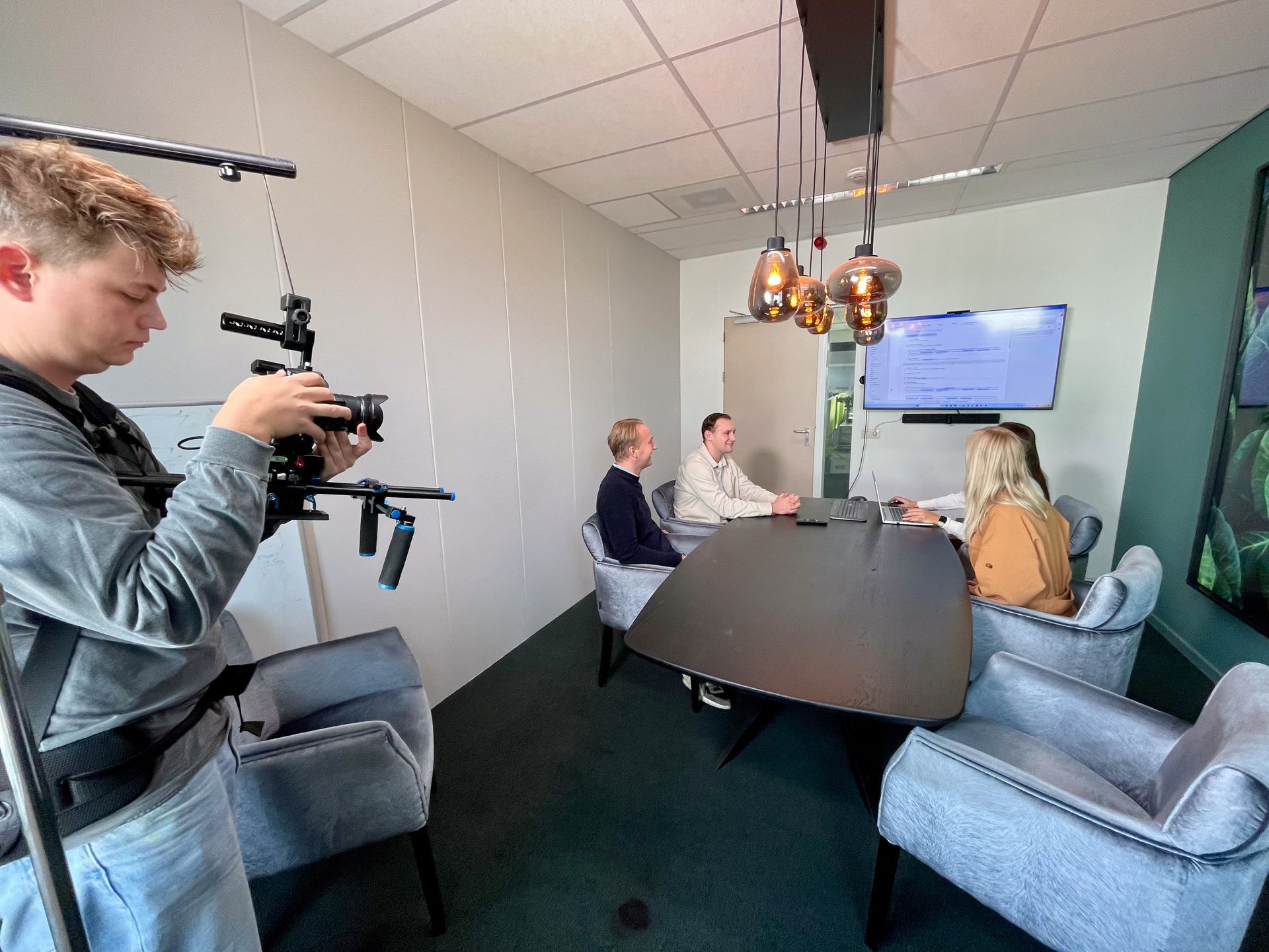 Iemand filmt een zakelijke bijeenkomst. Vier mensen zitten rond een tafel. Moderne kantooromgeving met videoscherm.