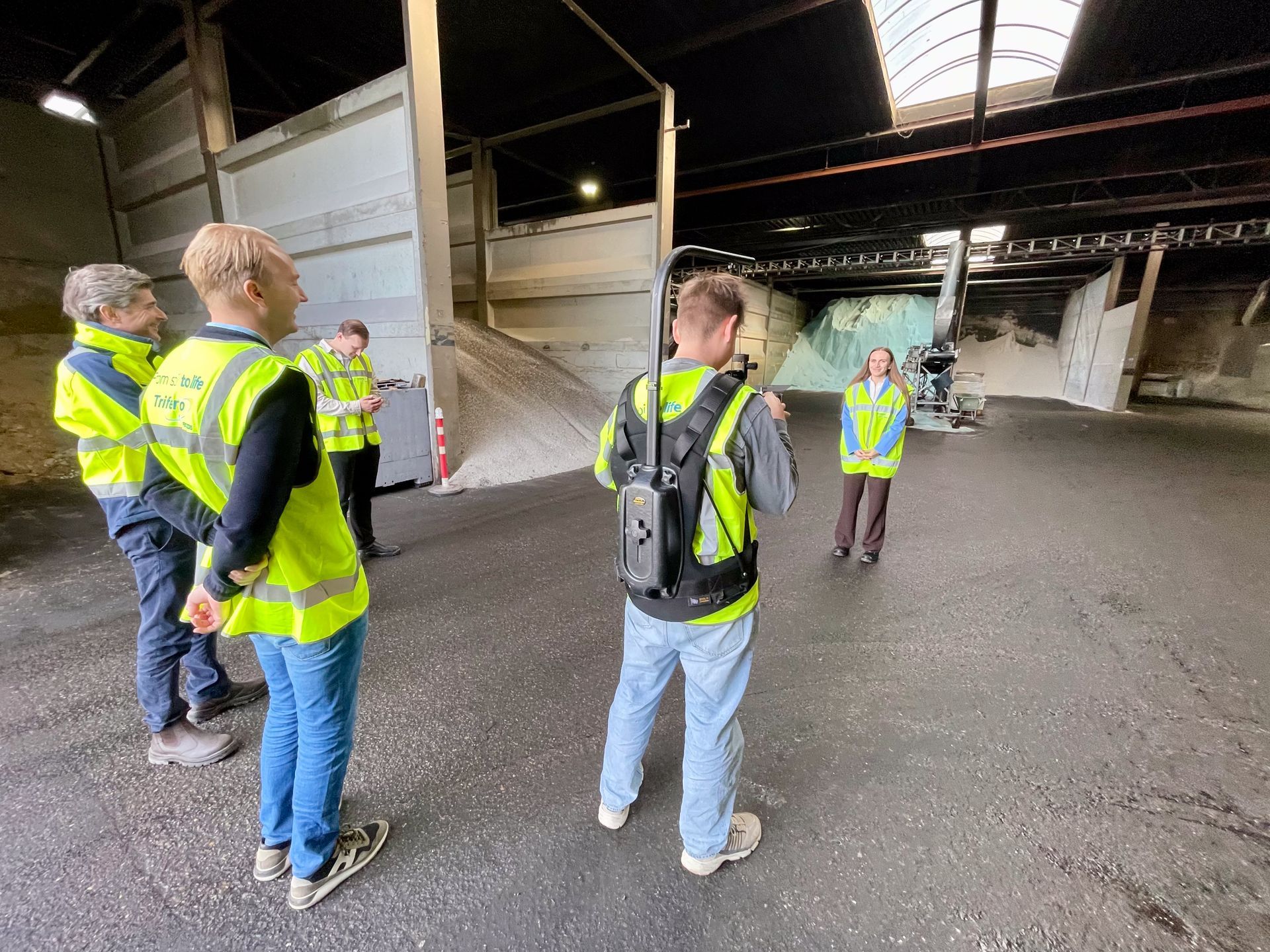 Mensen in veiligheidsvesten in een groot gebouw, mogelijk een magazijn. Ze kijken naar iets.