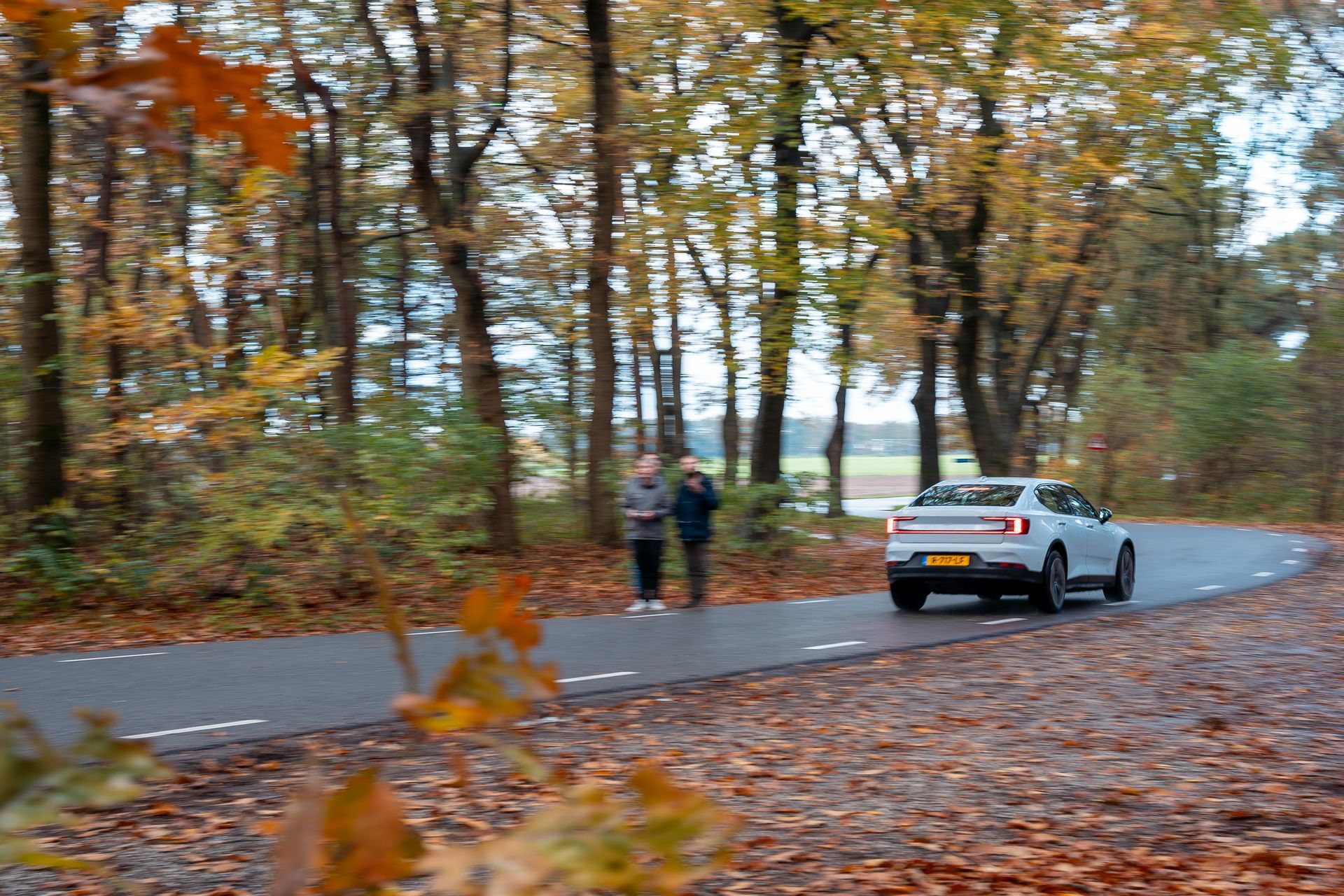 Een witte auto rijdt over een bochtige weg door een kleurrijk herfstbos; twee mensen staan ​​er vlakbij.