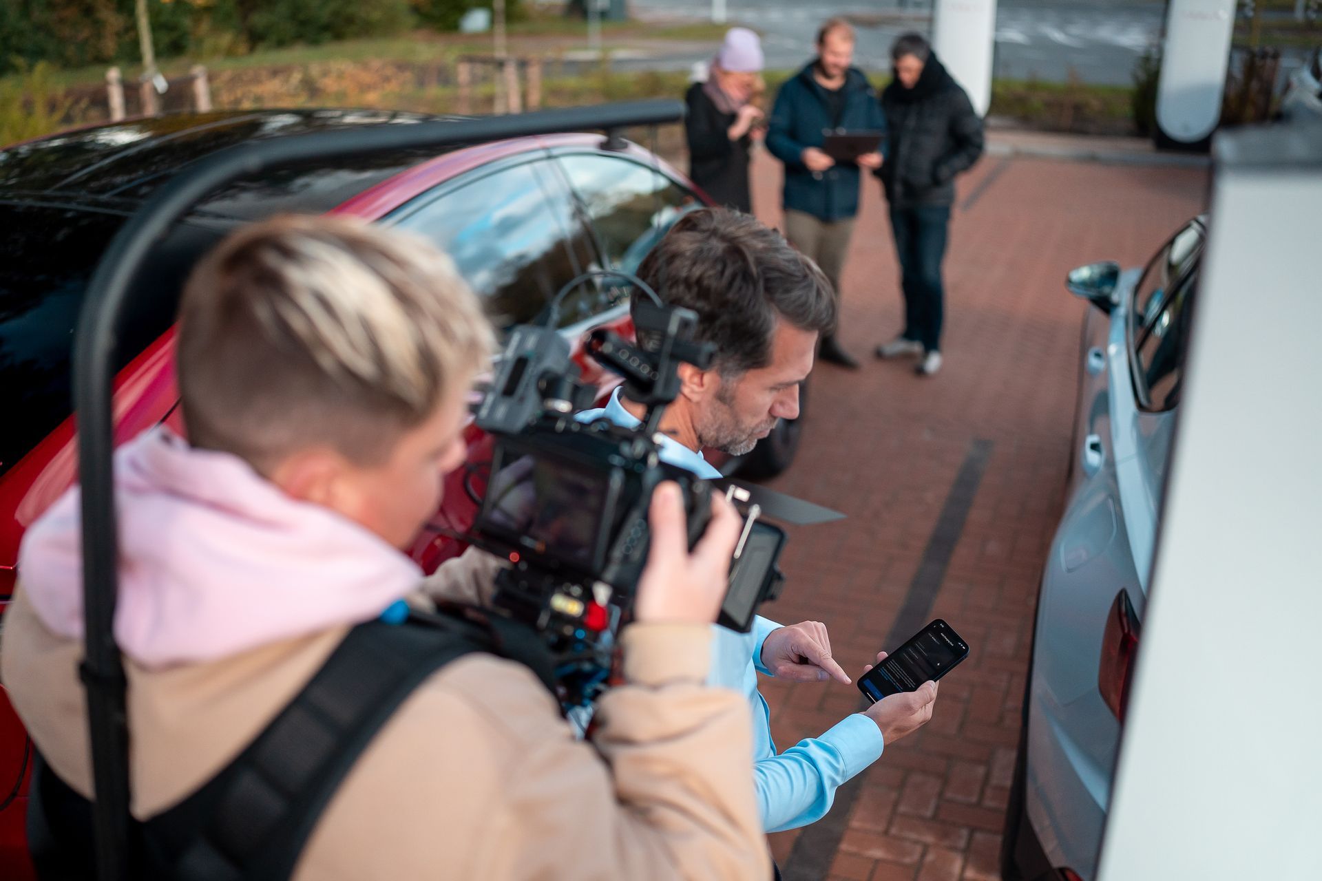 Iemand filmt een elektrische auto die aan het opladen is, met drie mensen op de achtergrond.