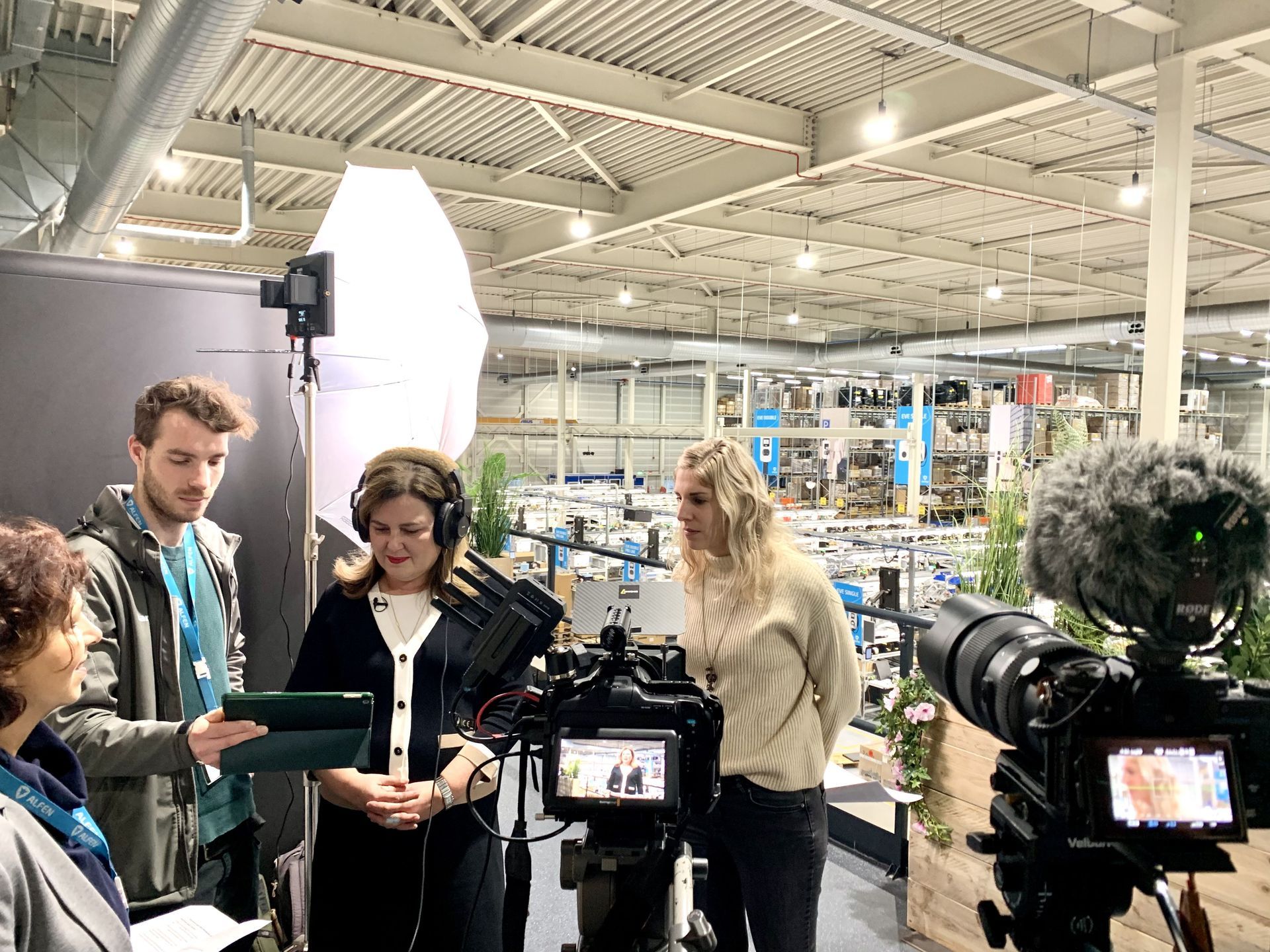 Een cameraploeg filmt in een groot magazijn. Drie mensen staan met microfoons en lampen bij de camera.