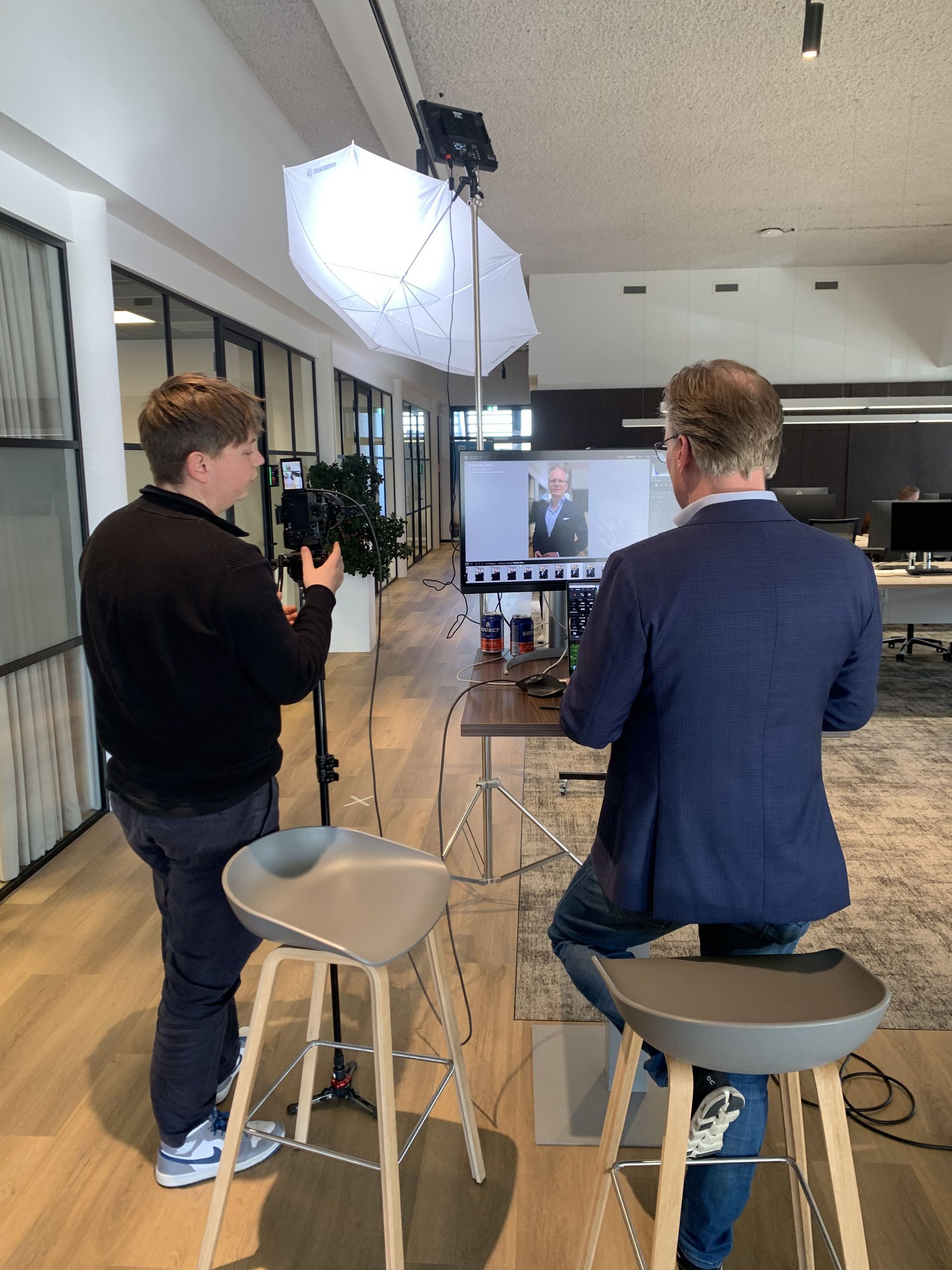 Iemand filmt iemand anders, die op een krukje zit, in een modern kantoor. Verlichting en een computerscherm zijn zichtbaar.