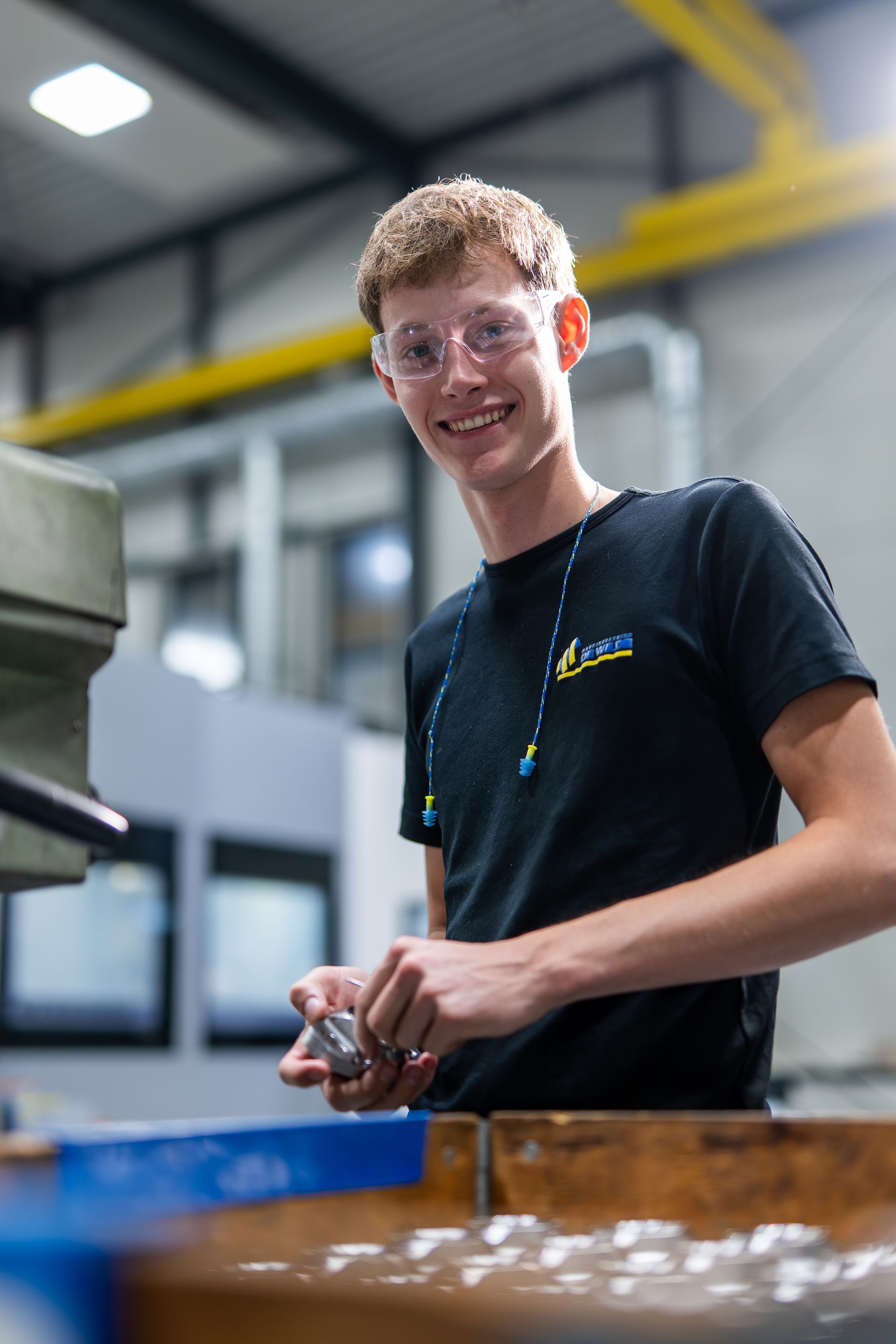 Een persoon met een veiligheidsbril lacht tijdens het werk in een productieomgeving.