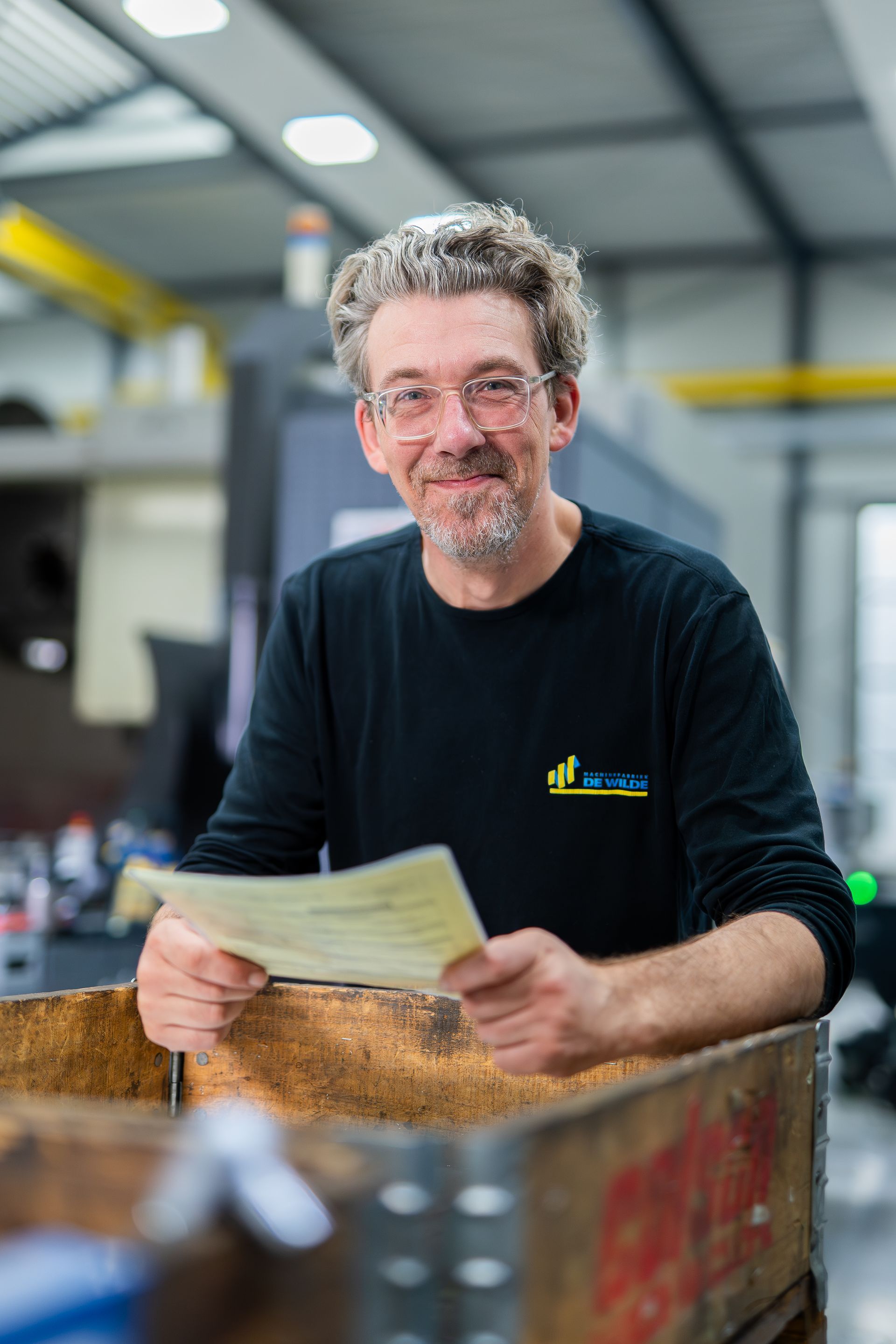 Een man met een bril, die een vel papier vasthoudt, glimlacht in een werkplaats. Hij draagt ​​een zwart shirt.