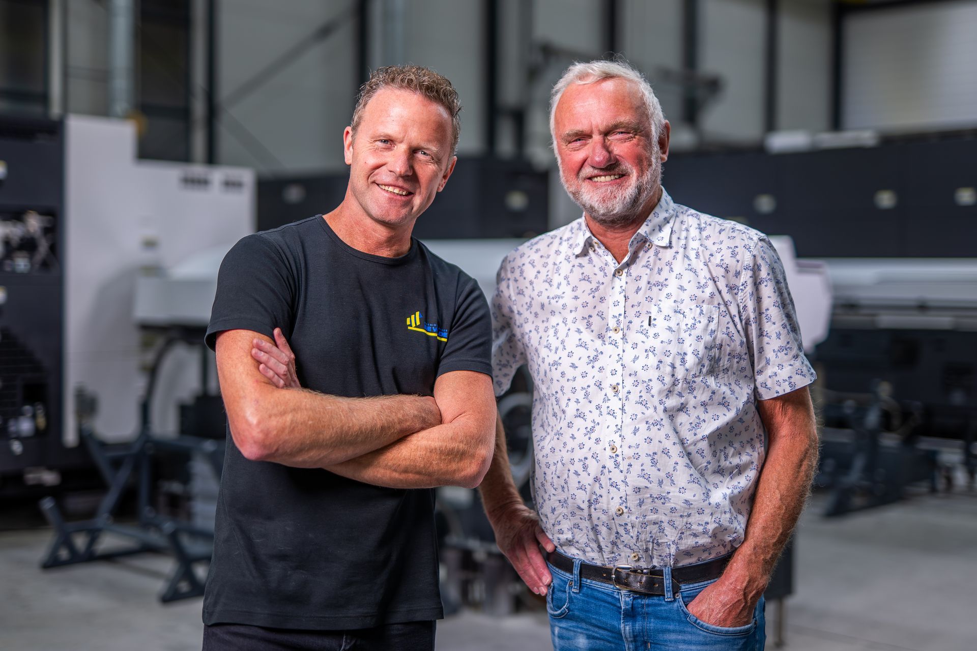 Twee lachende mannen poseren in een drukkerij. De ene man heeft zijn armen over elkaar, de andere heeft zijn handen in zijn zakken.