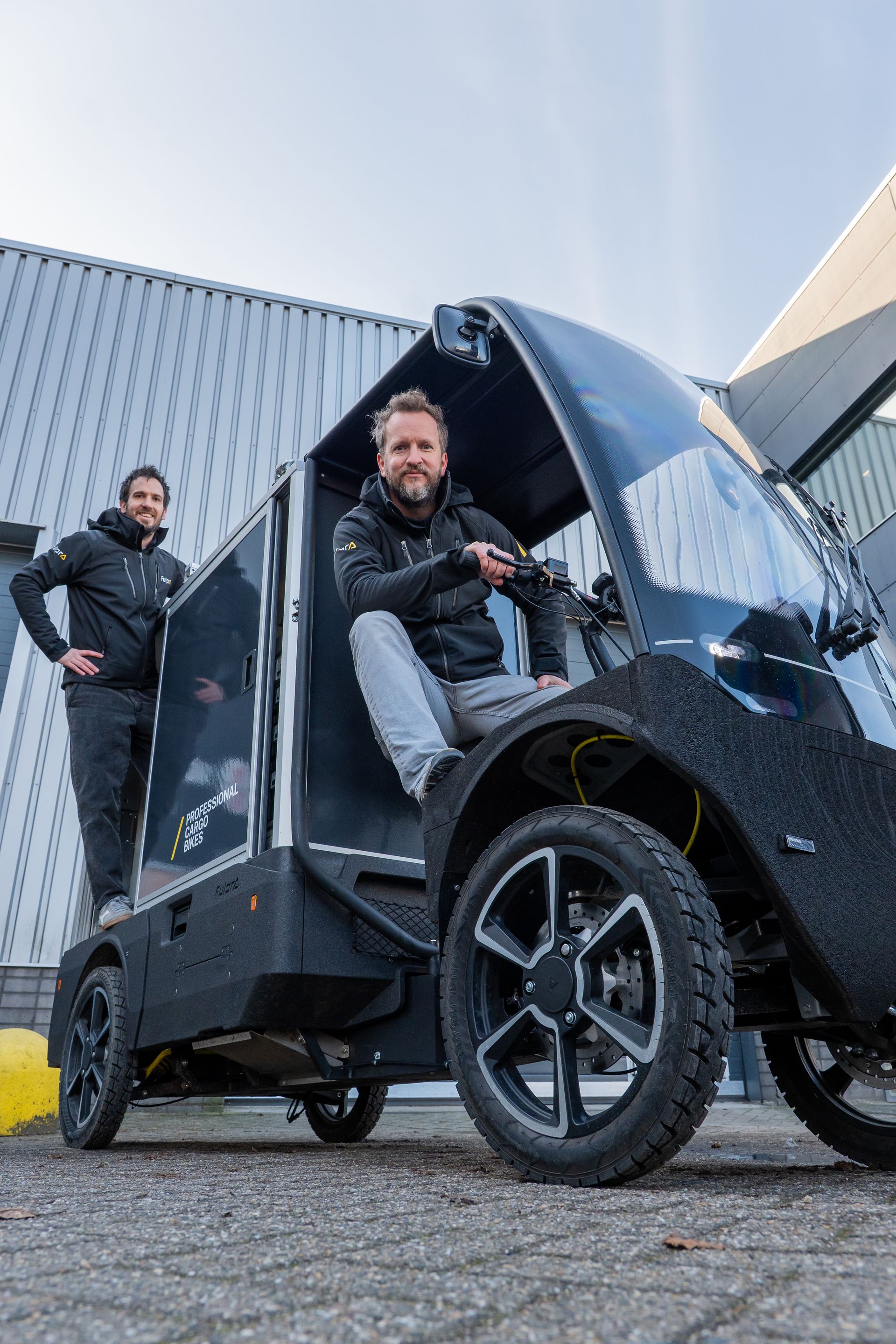 Twee mannen bij een zwarte elektrische bedrijfswagen. Eén zit achter het stuur, de ander staat bij de deur; beiden glimlachen.