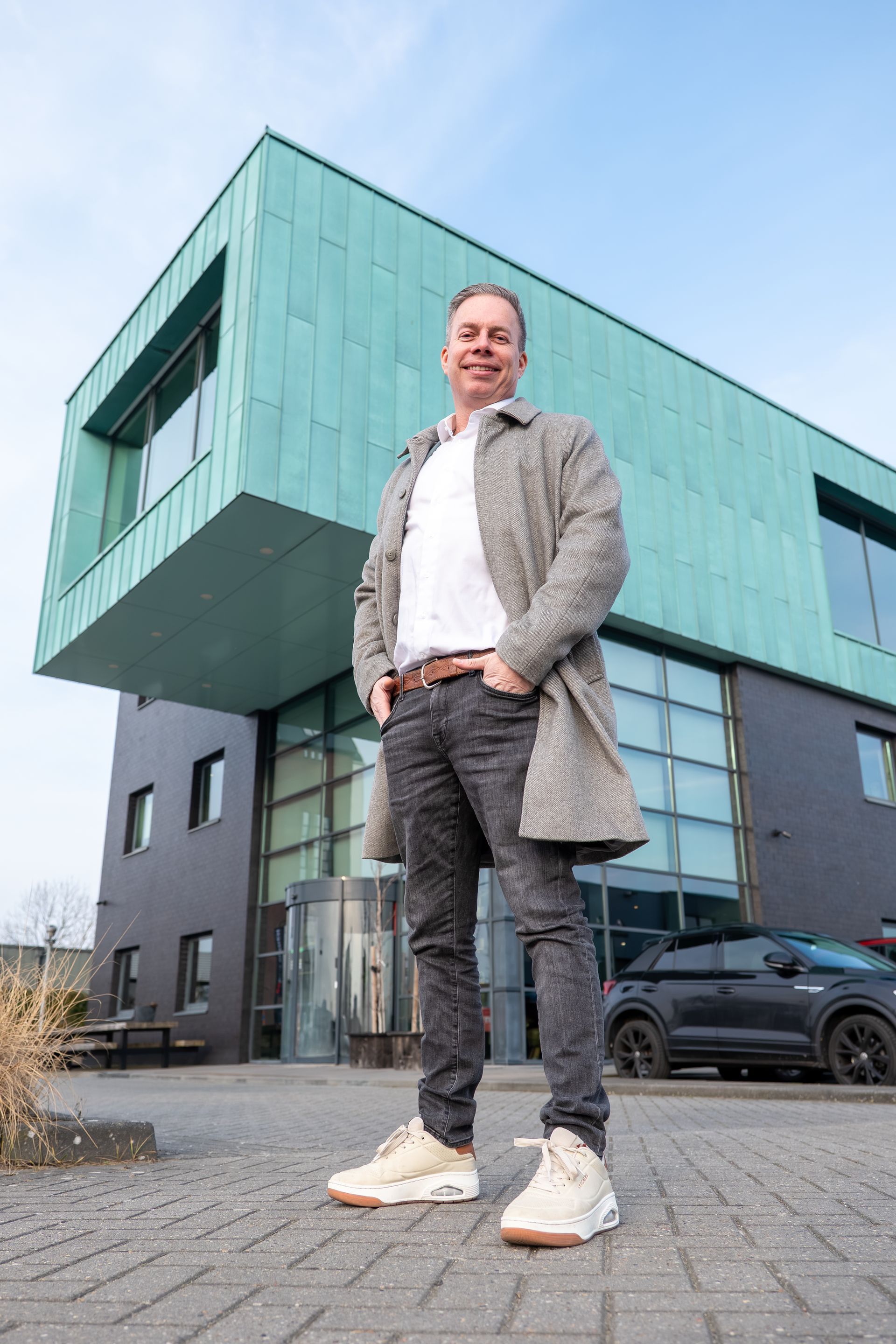 Een man in een jas staat voor een modern gebouw met een blauwgroene gevel.
