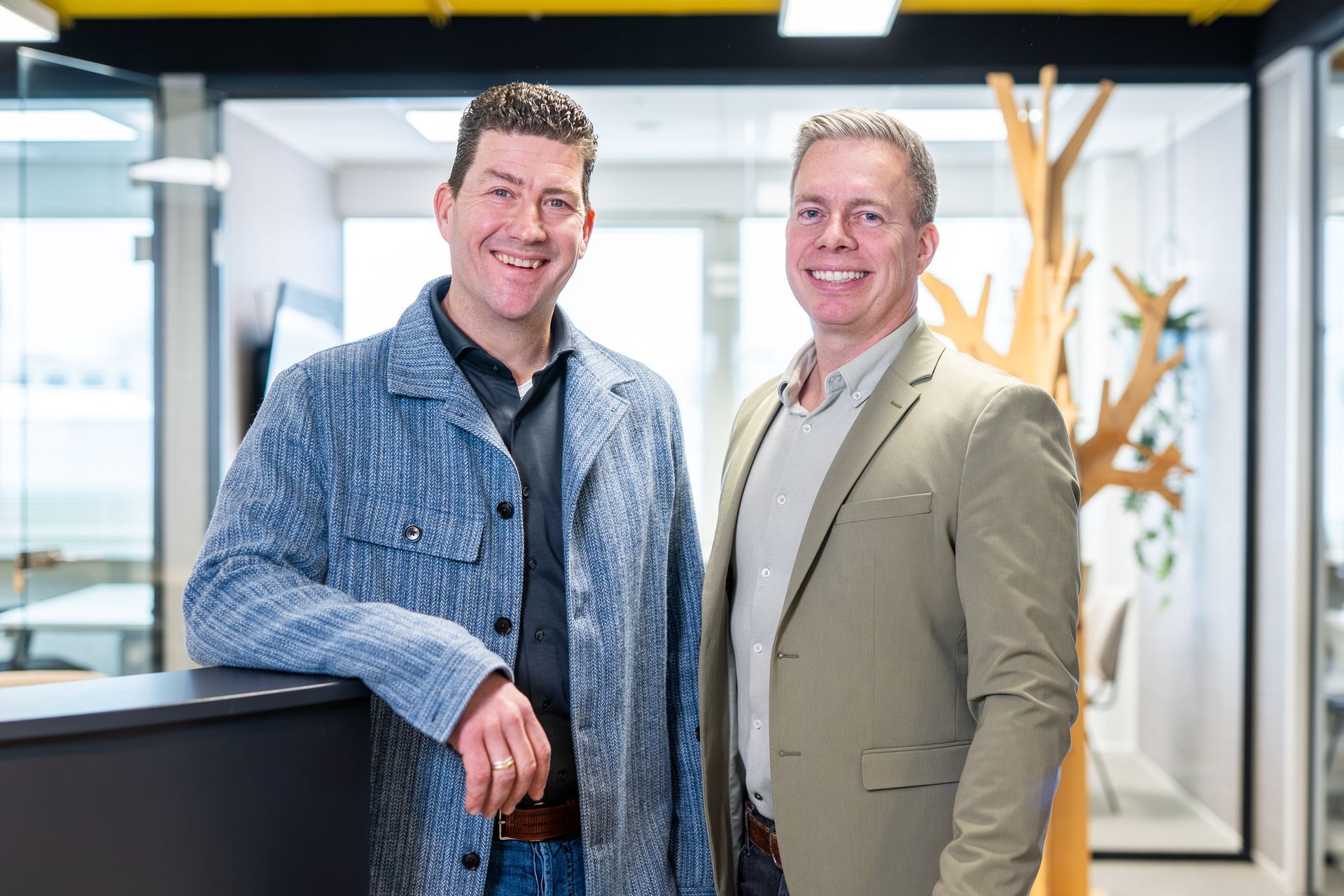 Twee glimlachende mannen, de een in een spijkerjasje, de ander in een beige blazer, staan ​​in een kantoor.