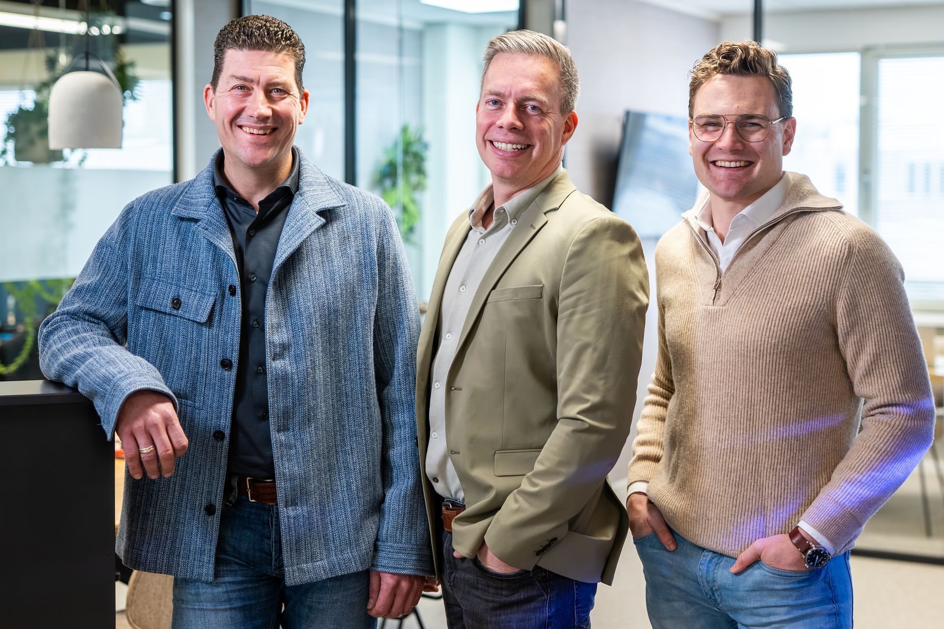 Drie mannen staan ​​binnen te glimlachen. De middelste man draagt ​​een groene blazer, de andere twee een casual jasje.