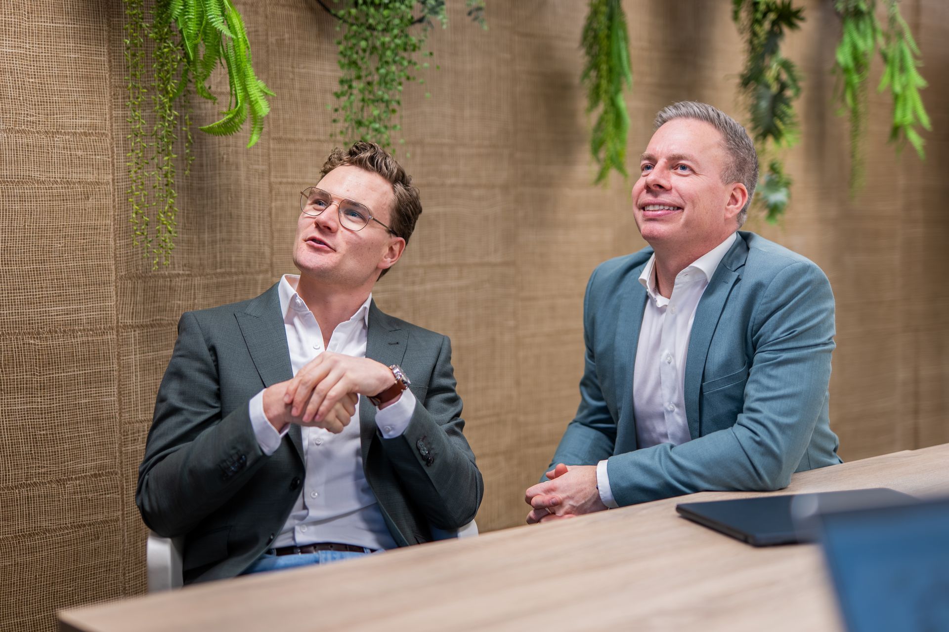 Twee mannen in pak zitten aan een tafel in een modern kantoor en kijken omhoog.
