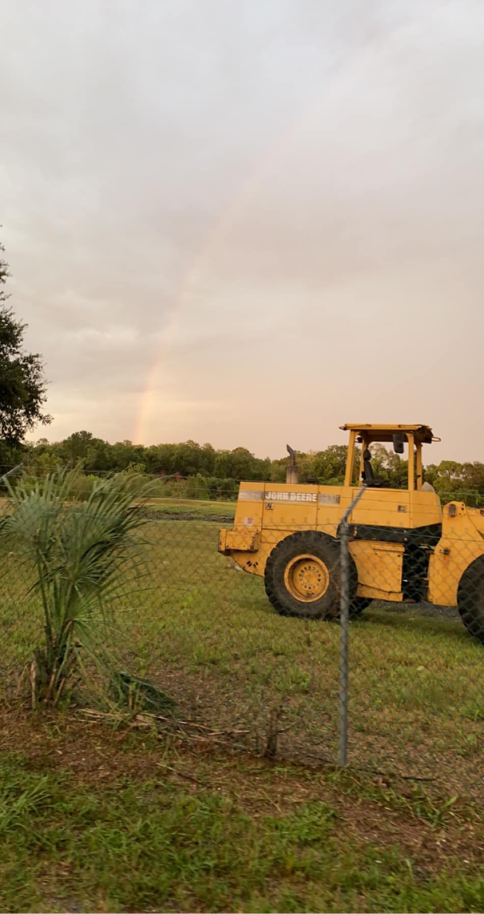Yellow construction vehicle on grass field with a small rainbow against an overcast sky.