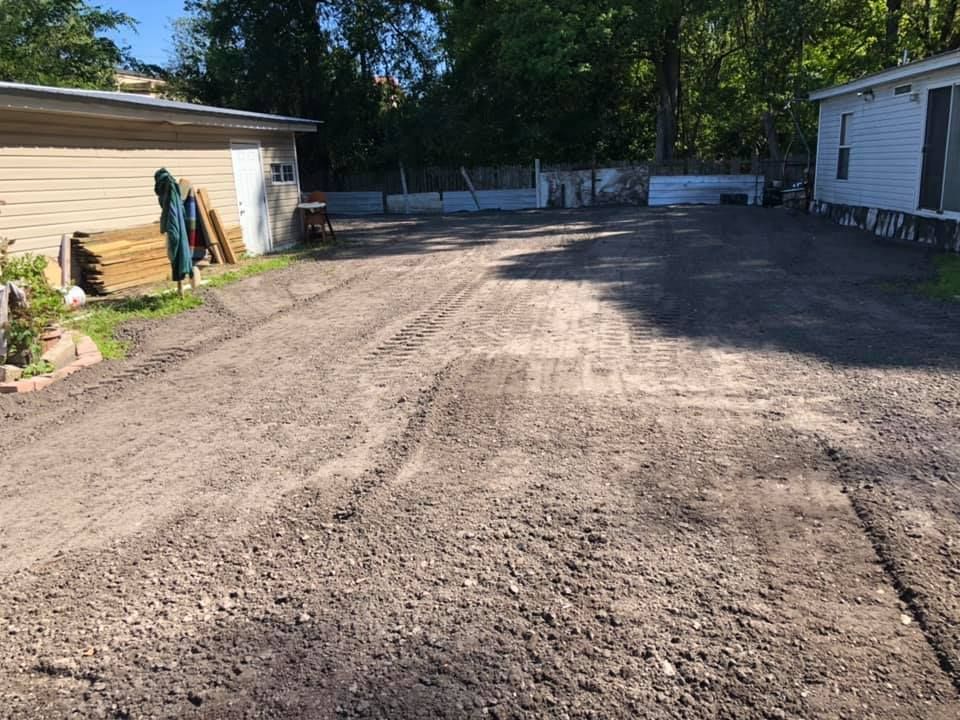 Gravel driveway in front of a white garage and house; lumber leaning against garage, sunny day.