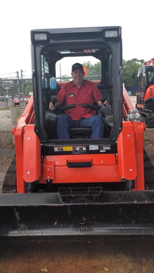 Man seated in the cab of an orange skid steer loader, smiling; outdoors.