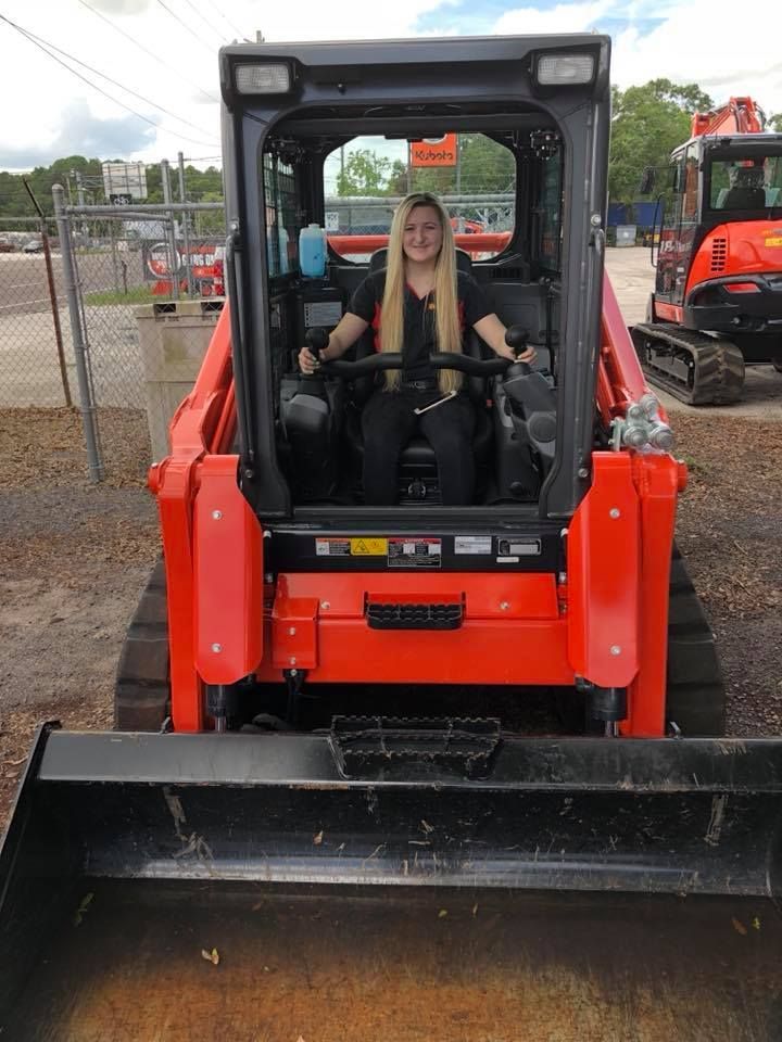 Woman operating an orange skid steer loader. She is sitting inside, smiling. Outdoor setting.