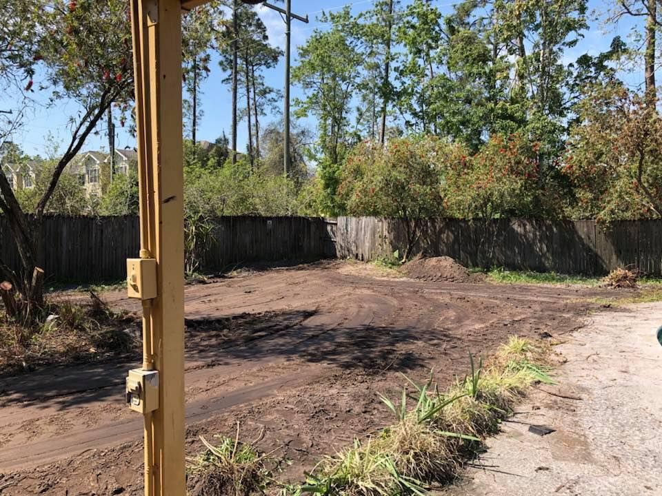 Dirt lot in front of a wooden fence, with green grass and trees in the background. A yellow post is in the foreground.