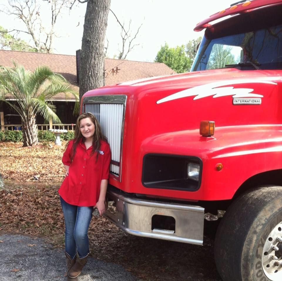 Woman in red shirt and jeans leans on a red semi-truck. Outdoors, house in background.