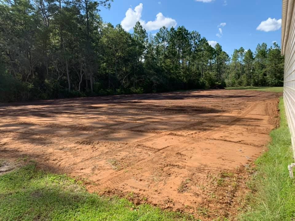 A cleared dirt lot, brown soil, next to a building and trees under a blue sky.