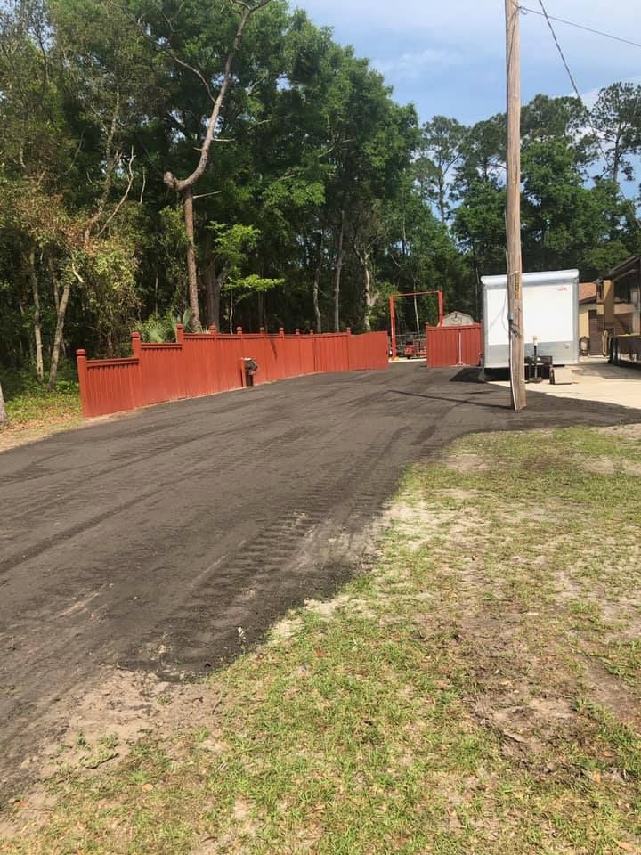 Newly paved asphalt driveway leading toward a red fence and trees, with a utility pole and a white trailer on the side.