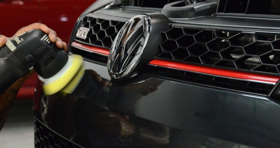 Person polishing a black car bumper with a buffer, near the VW emblem and red stripe.
