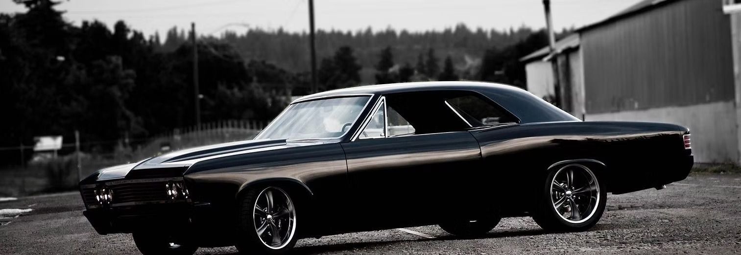 Black classic car parked next to a building and trees in a black and white scene.