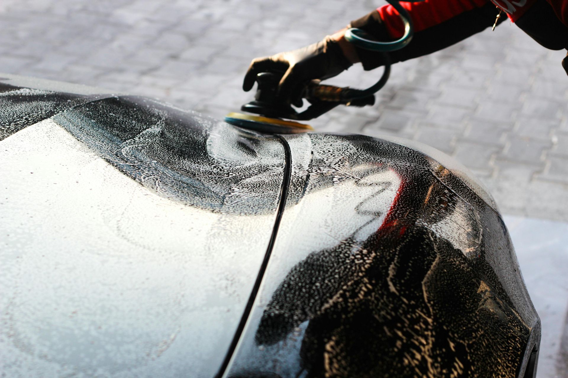 A person polishing a car's dark surface with a buffer, creating swirls of soap.