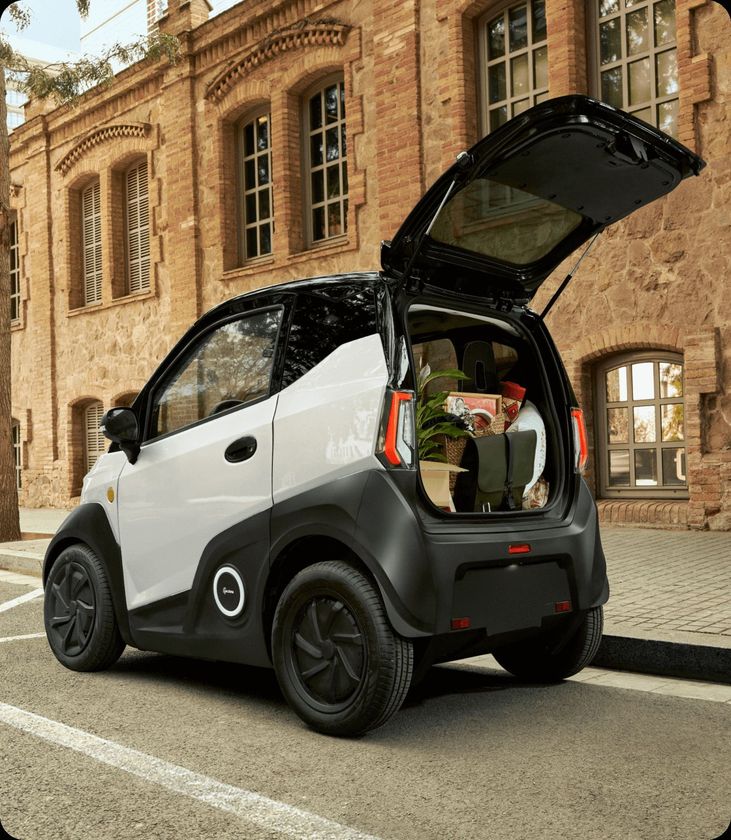 Una piccola automobile elettrica con il bagagliaio aperto è parcheggiata davanti a un edificio in mattoni.