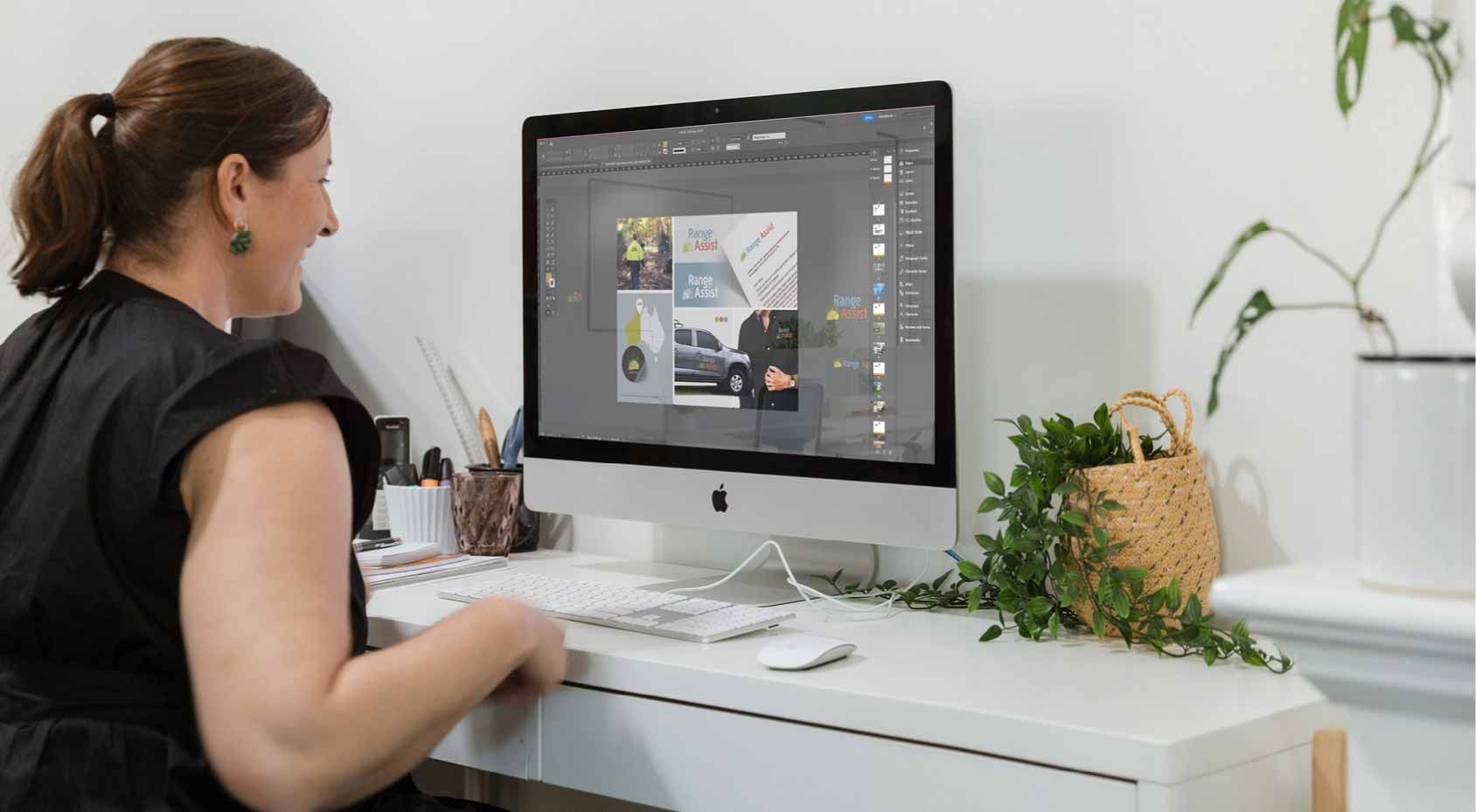 Woman working at a desk on an iMac in a bright home office, with plants and papers nearby.