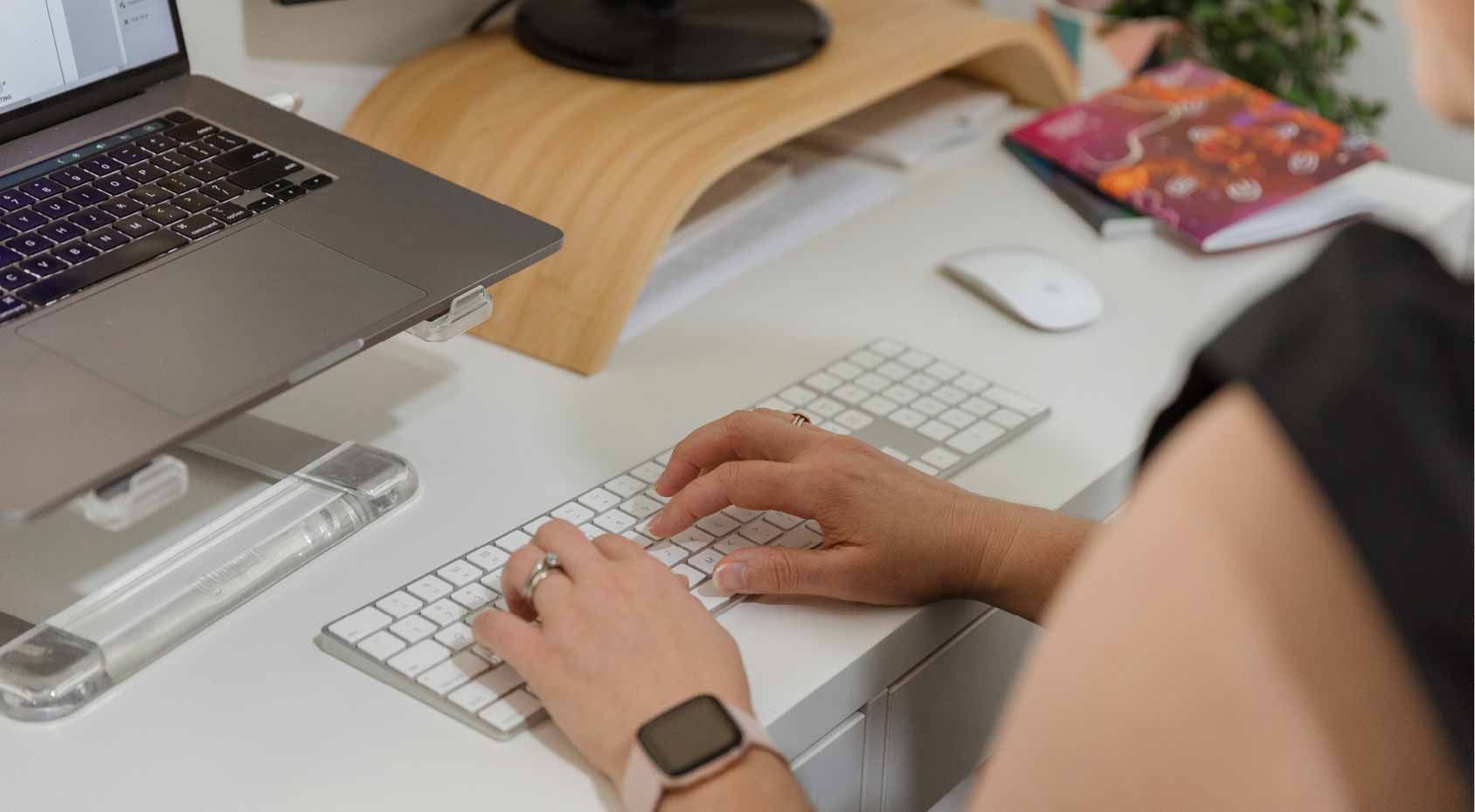 Person typing on a white keyboard at a desk beside a laptop and mouse.