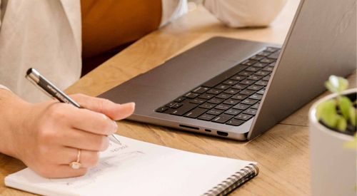 Hand writing on paper beside an open laptop on a wooden desk