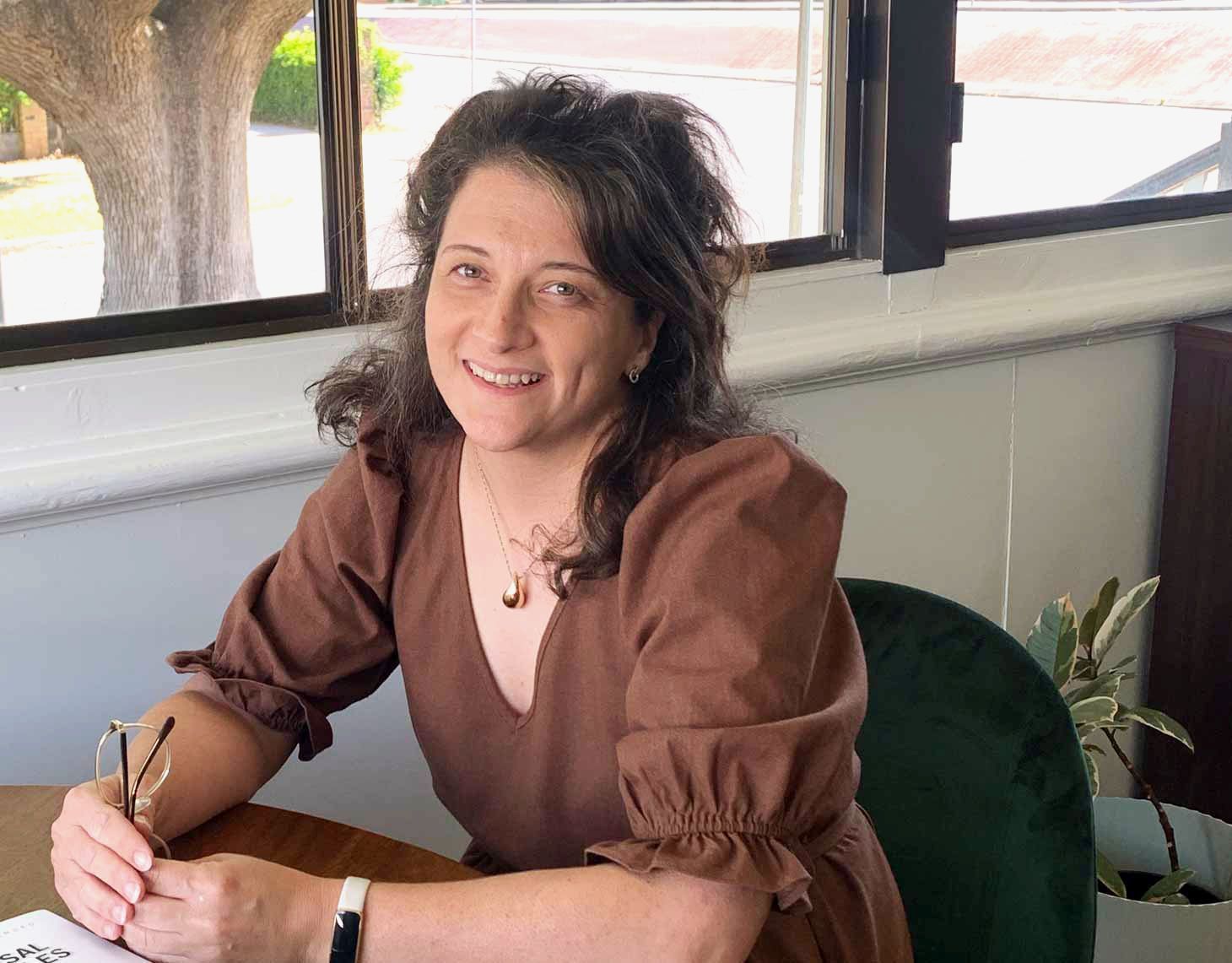 Woman smiling at a desk by a window, holding a pen and wearing a brown dress.