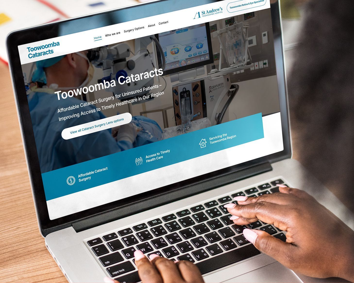 Hands typing on a laptop showing the Toowoomba Cataracts website with a teal header and navigation menu.