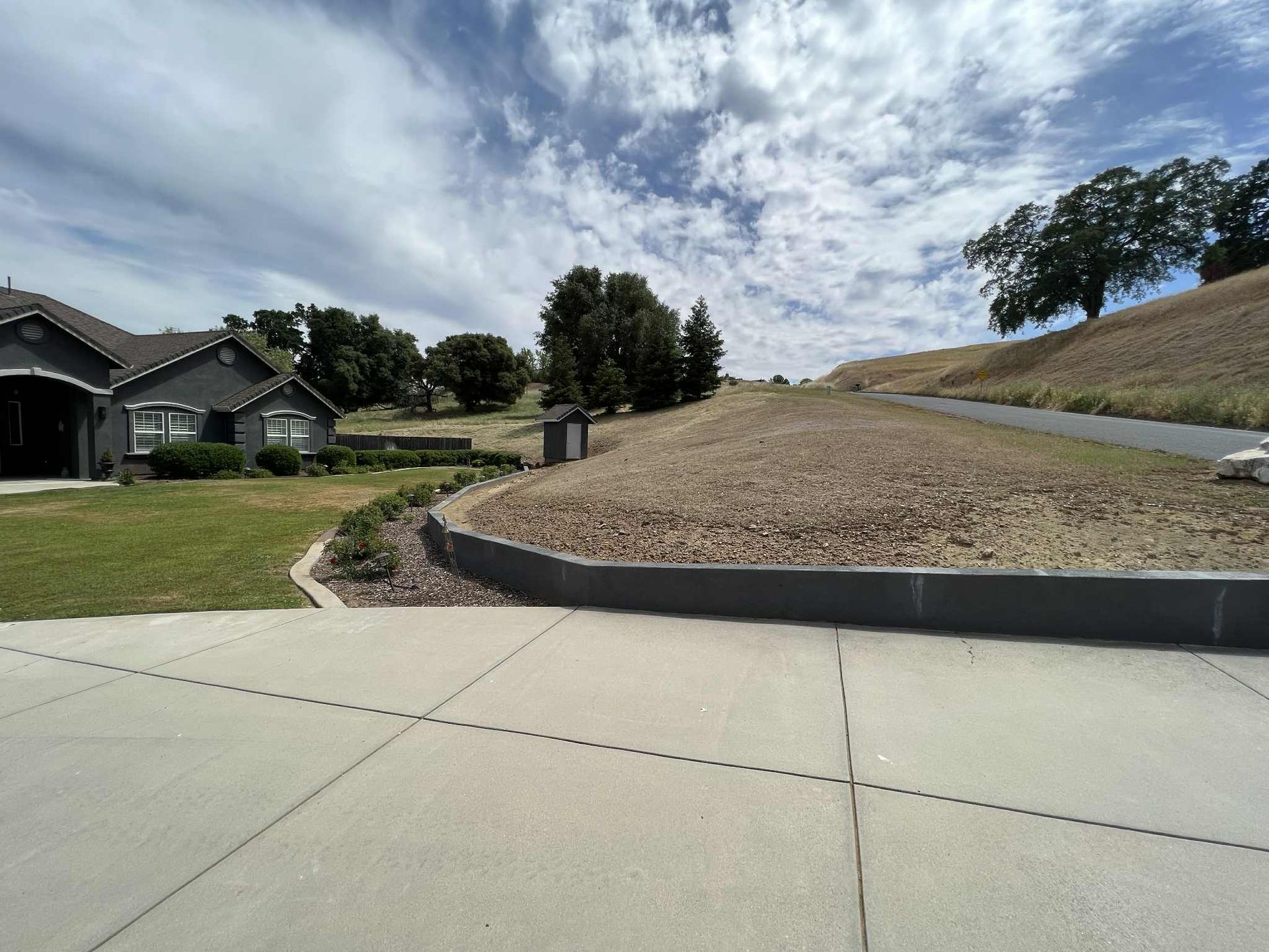 A house with a driveway next to a dry, grassy hill under a cloudy sky.