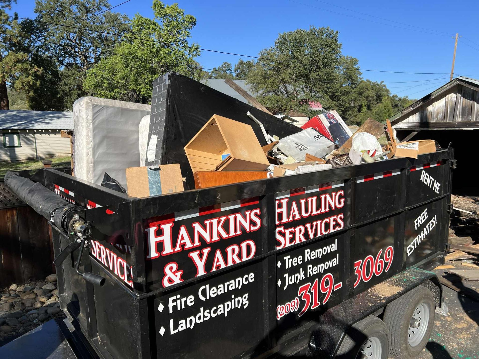 A black dumpster filled with trash from Hankins & Yard Hauling Services, outdoors, under a blue sky.